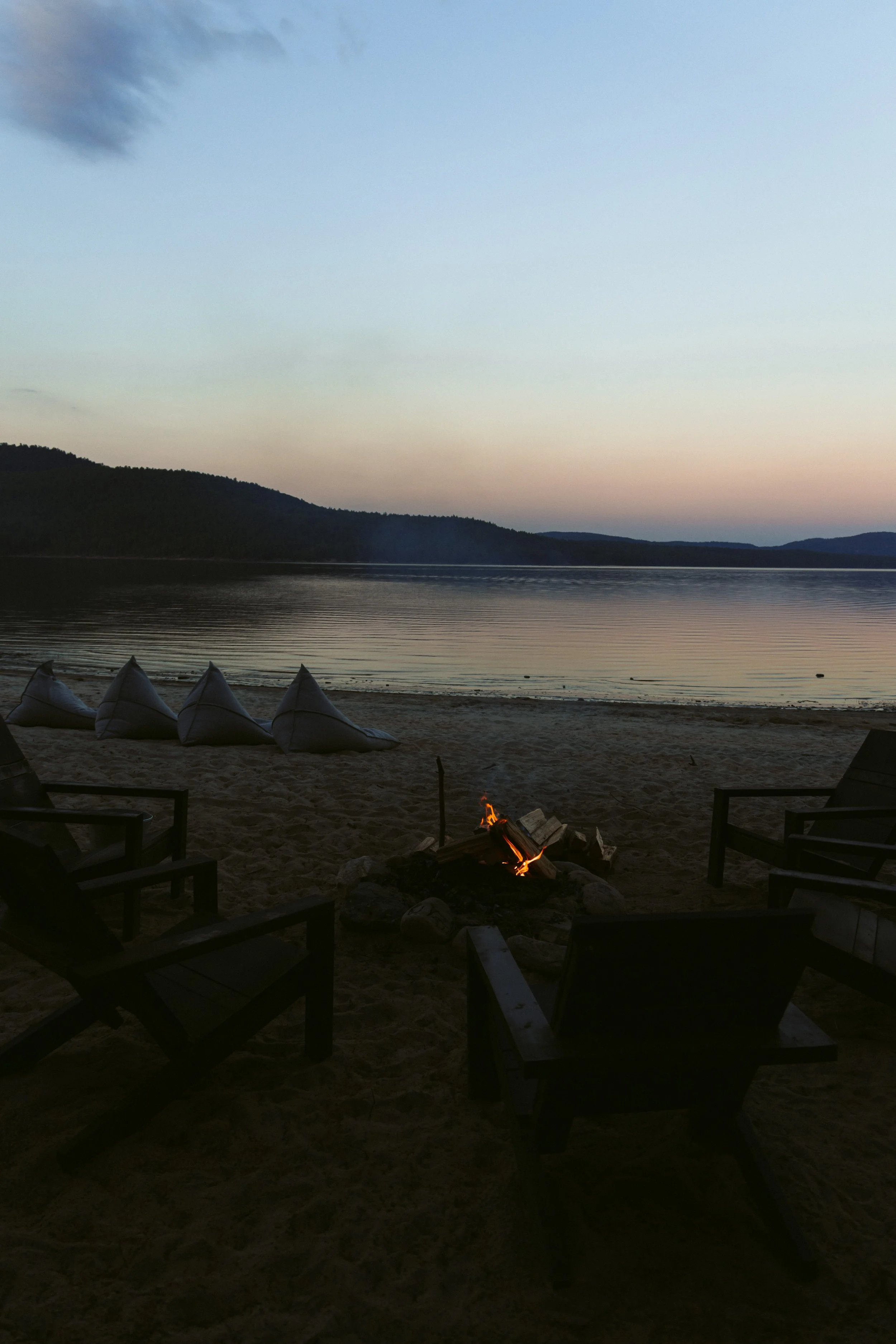 Beach at sunset with Adirondack chairs and a small campfire, kayaks on the sand, calm water, and mountains in the distance.