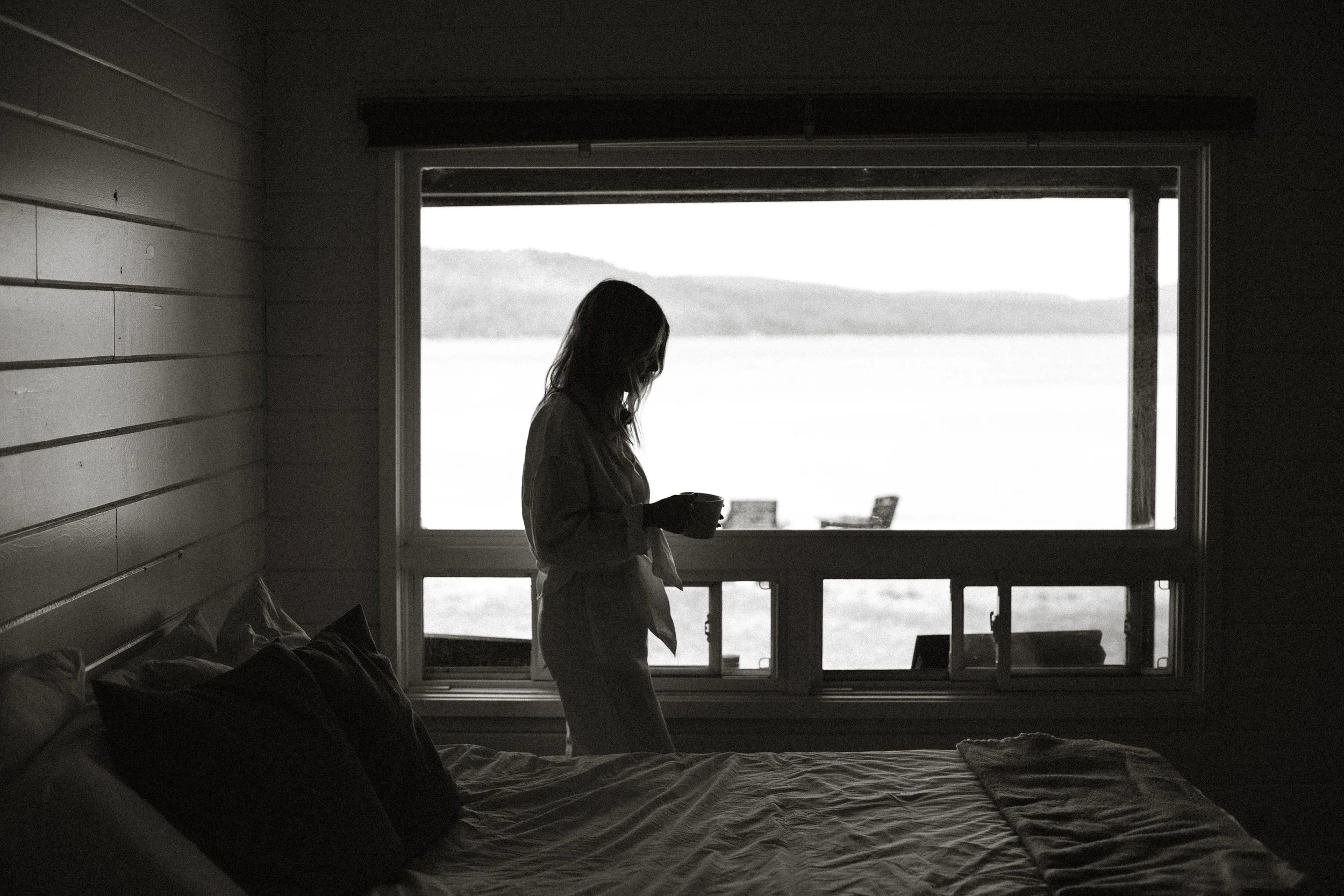 A woman standing by a window holding a mug inside a dark bedroom with a view of water and hills outside.
