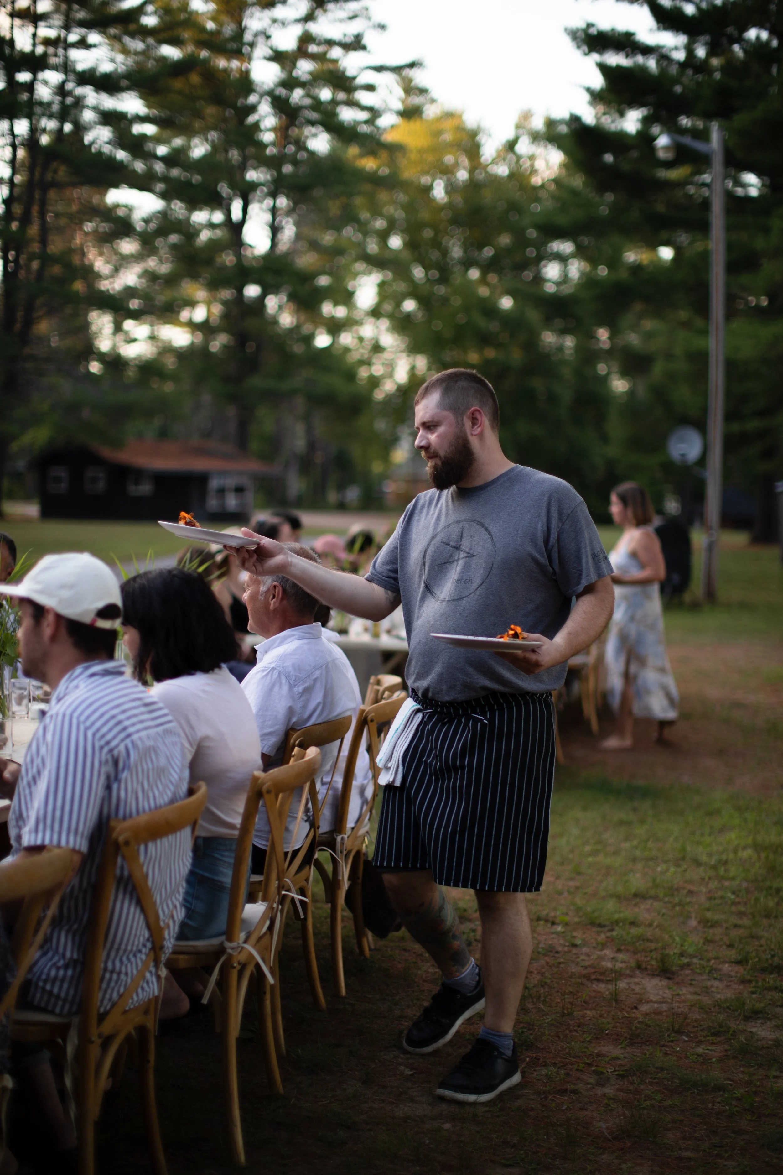 A man with a beard serving dinner at an outdoor gathering in a park, with guests seated at tables and colorful trees in the background during early evening.