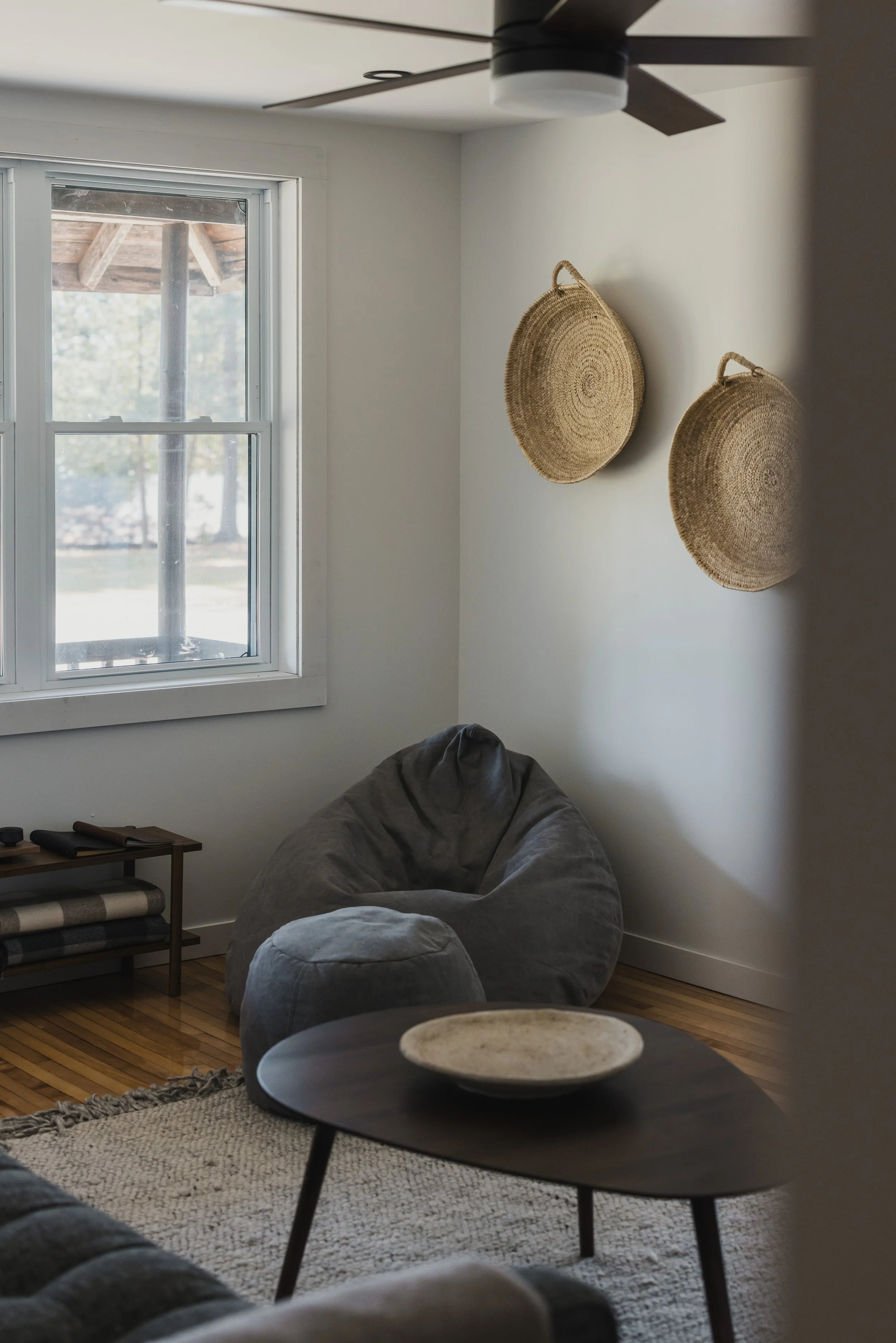 A cozy living room corner with a large window, a dark gray bean bag chair, a small round side table, woven wall baskets, and a ceiling fan.