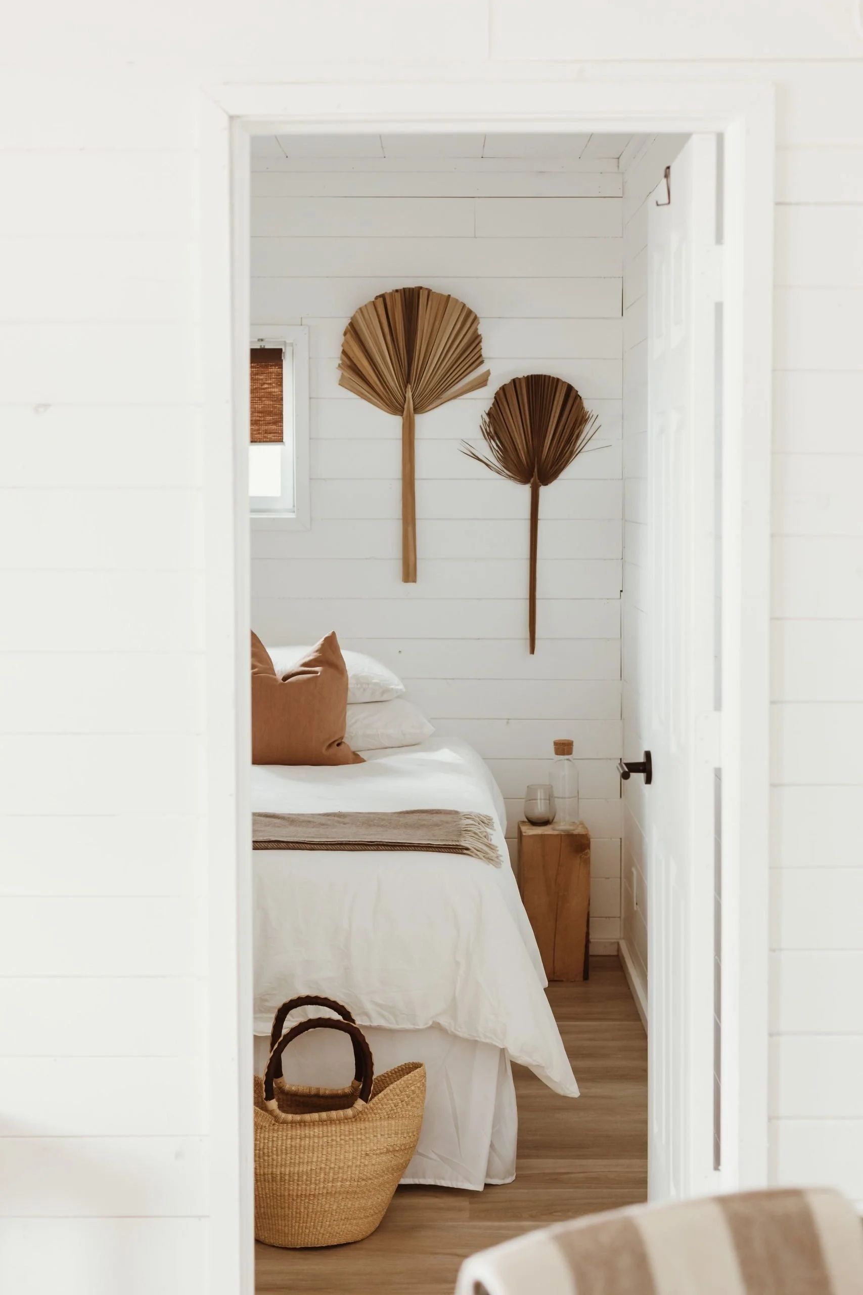 A cozy bedroom with white wooden walls, decorated with two large dried palm leaves mounted on the wall. The bed has white bedding, pillows, and a beige throw blanket. A small wooden side table holds a glass jar with a cork lid and a drinking glass. A woven basket with handles sits on the floor near the bed.