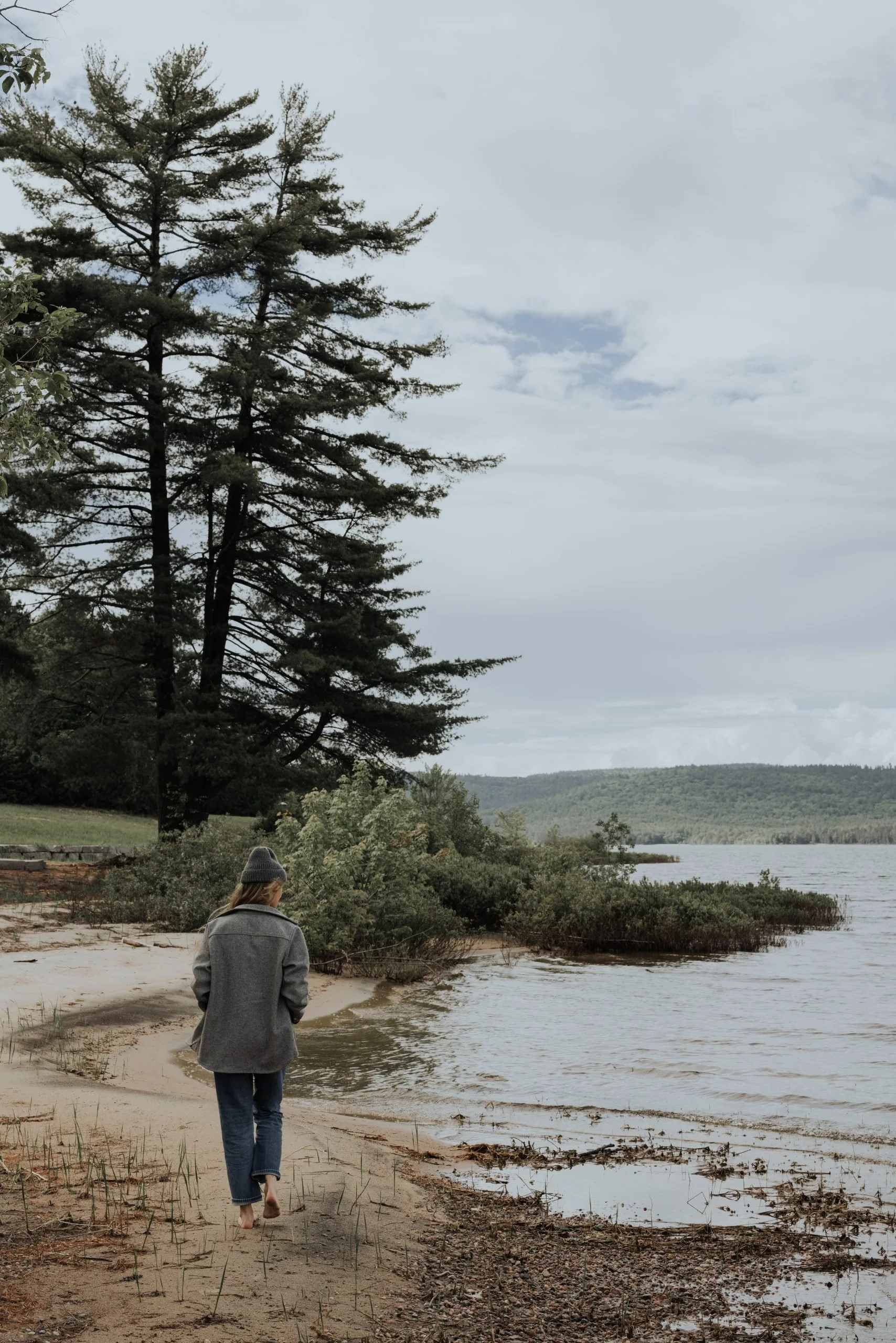 A person walking barefoot along a sandy lakeshore with trees and hills in the background on a cloudy day.
