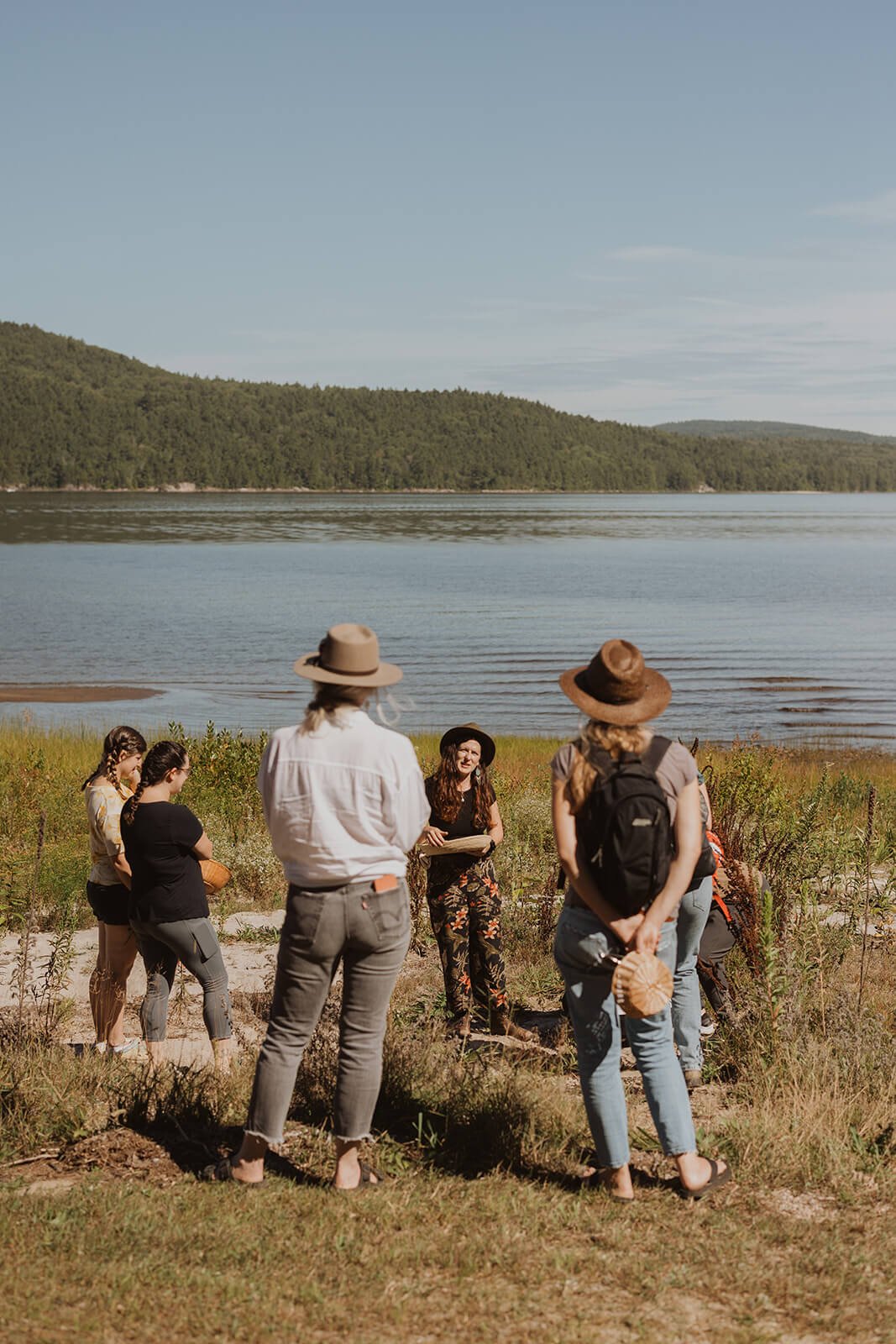 Group of people listening to a woman speaking near a lake with forested hills in the background.