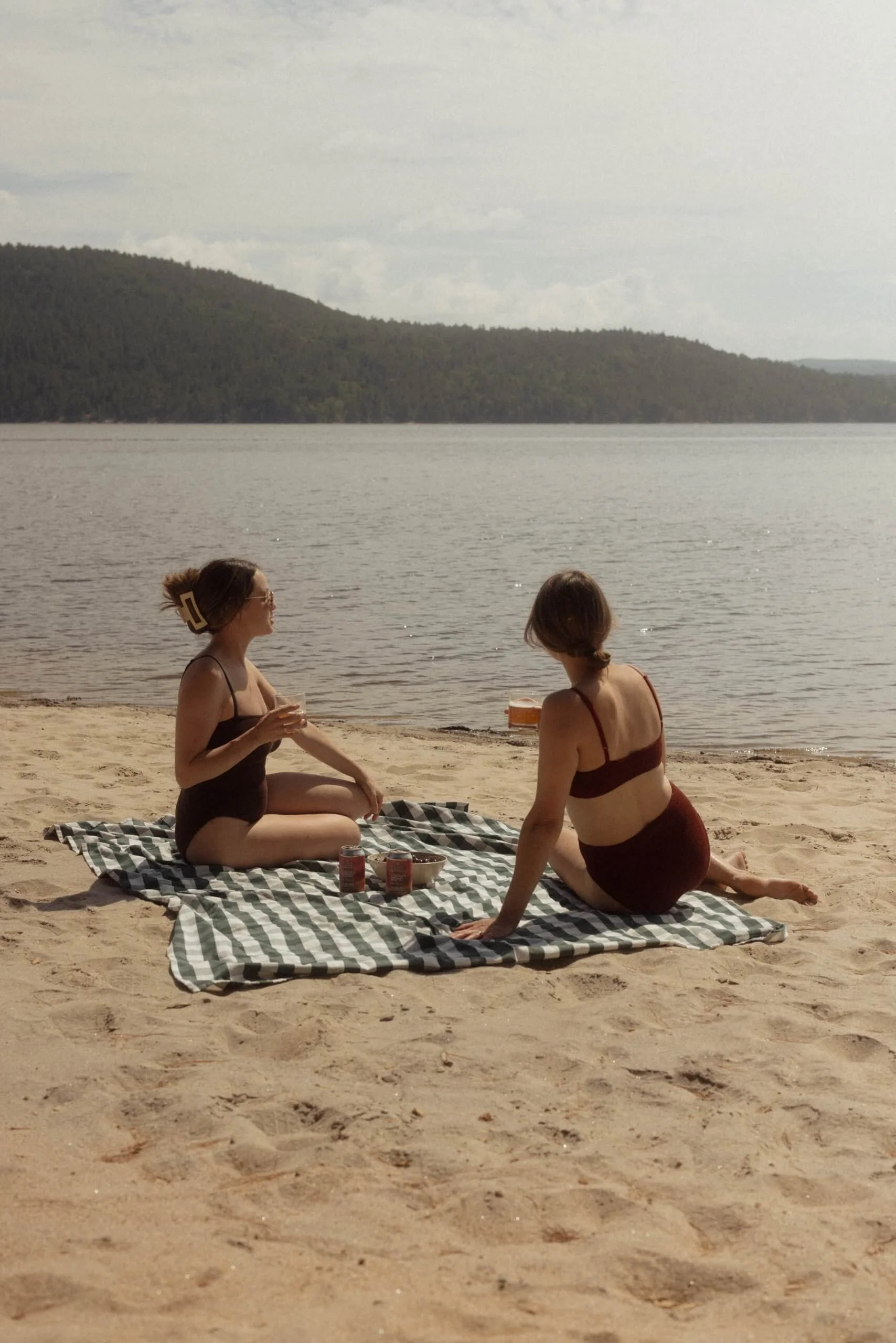 Two women in swimsuits sitting on a striped blanket on a sandy beach by a lake, with trees and hills in the background, enjoying drinks.