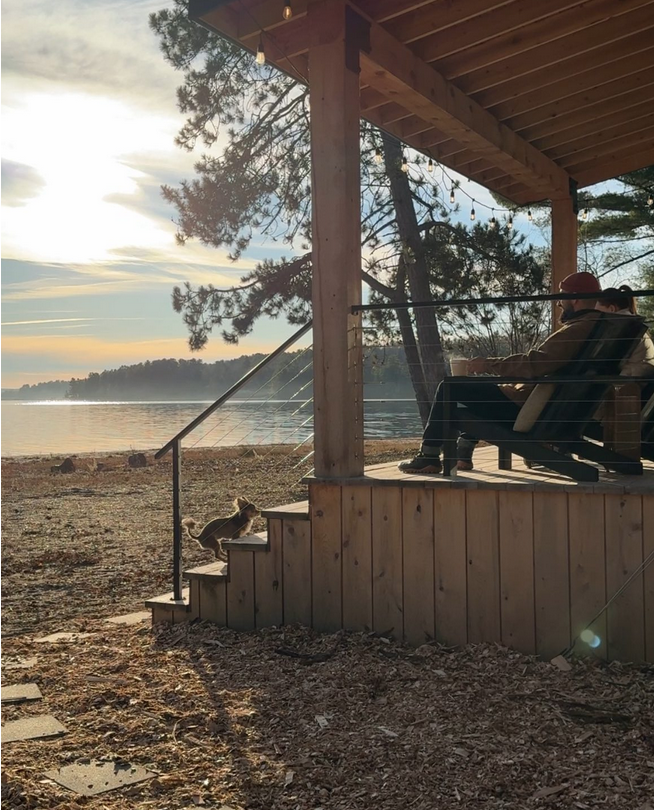 A person sitting on a porch near a lake at sunset, with a dog stepping down the stairs.