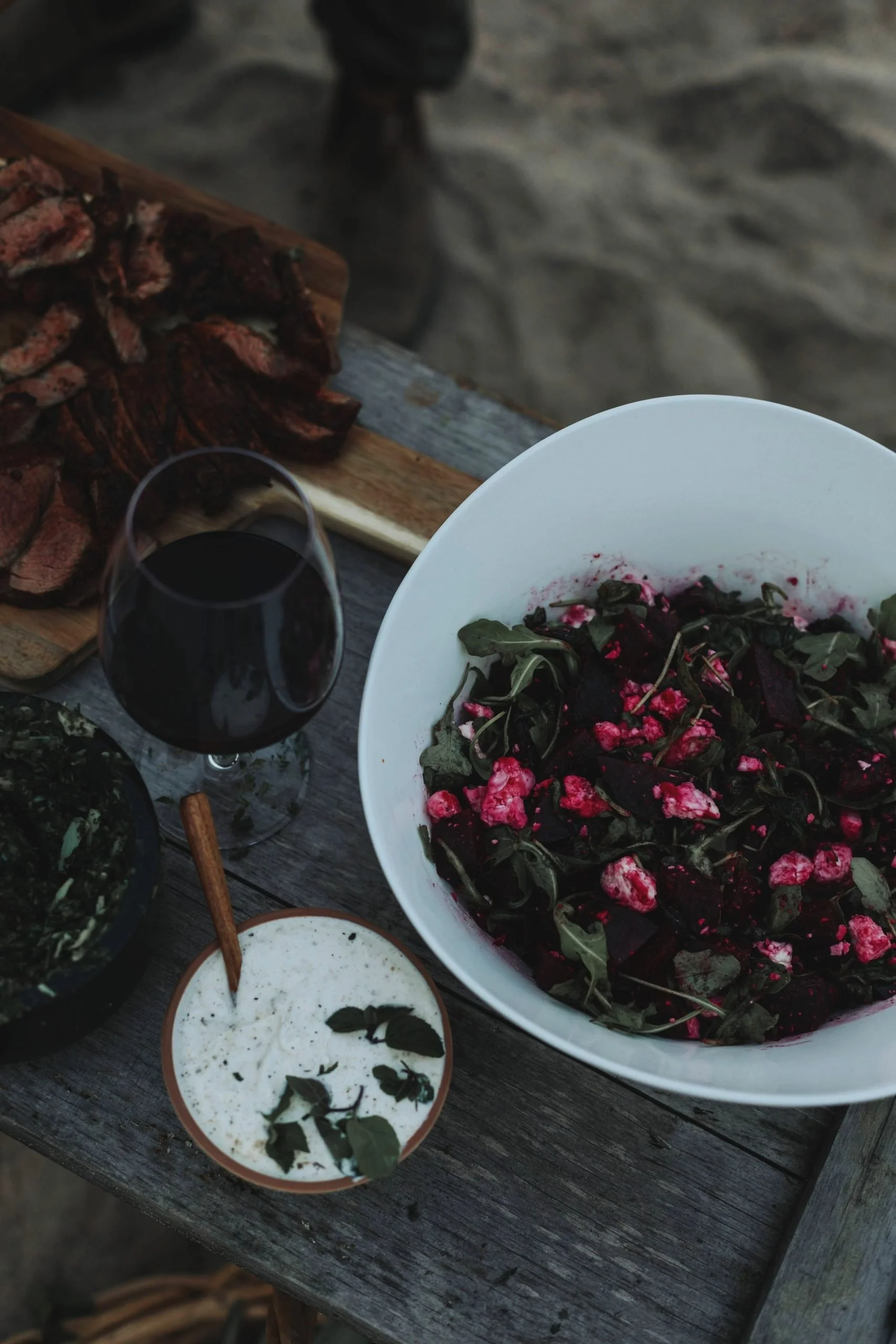 A rustic outdoor table with grilled meat, a glass of red wine, a veggie salad in a large white bowl, a bowl of creamy dip with herbs, and a small bowl of greens.