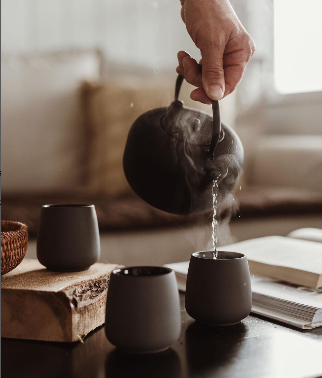 A hand pouring hot water from a brown teapot into one of three black ceramic cups on a wooden table.