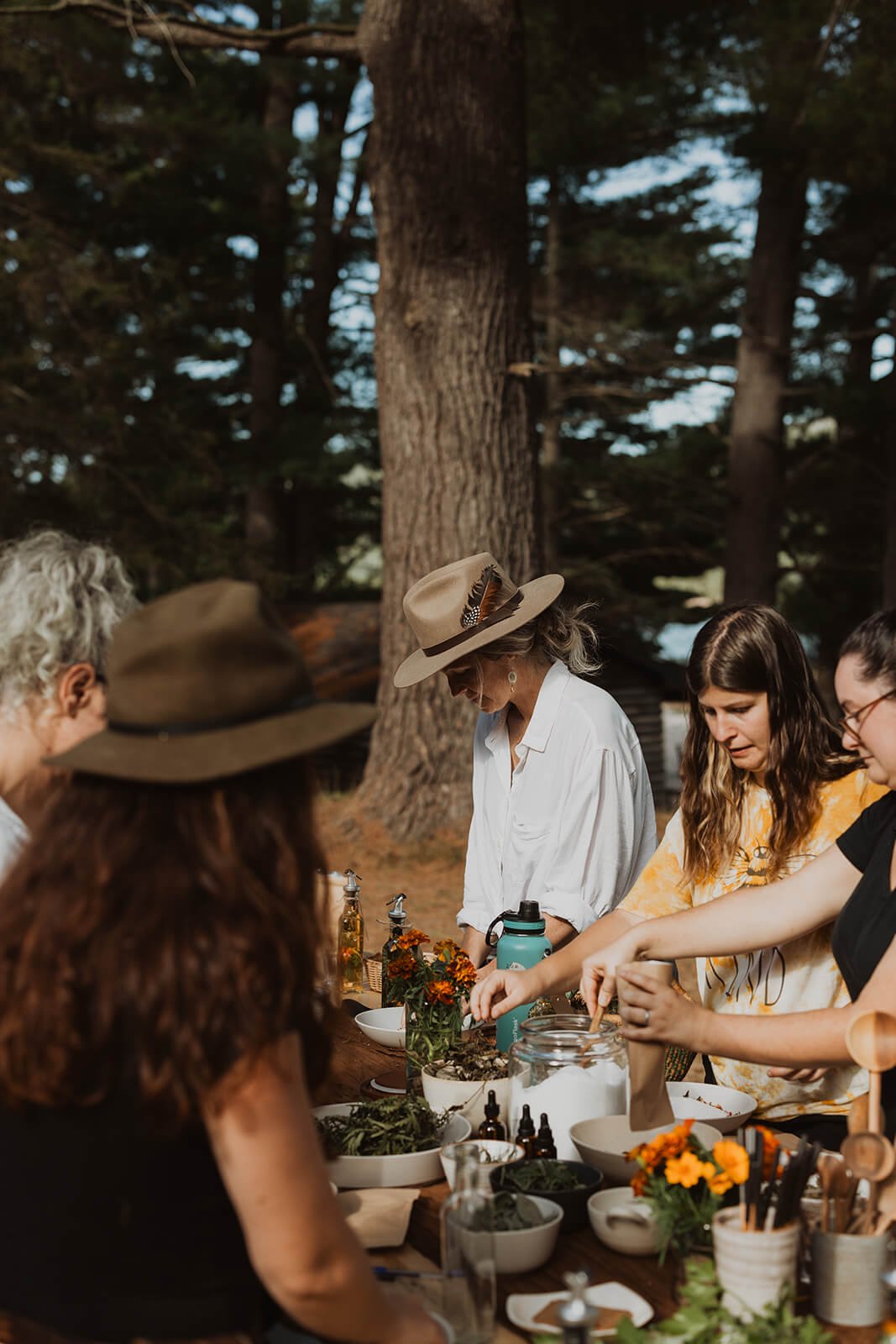 A group of people gathered around an outdoor table filled with bowls, bottles, and plants in a wooded area, engaging in a nature-related activity or craft.