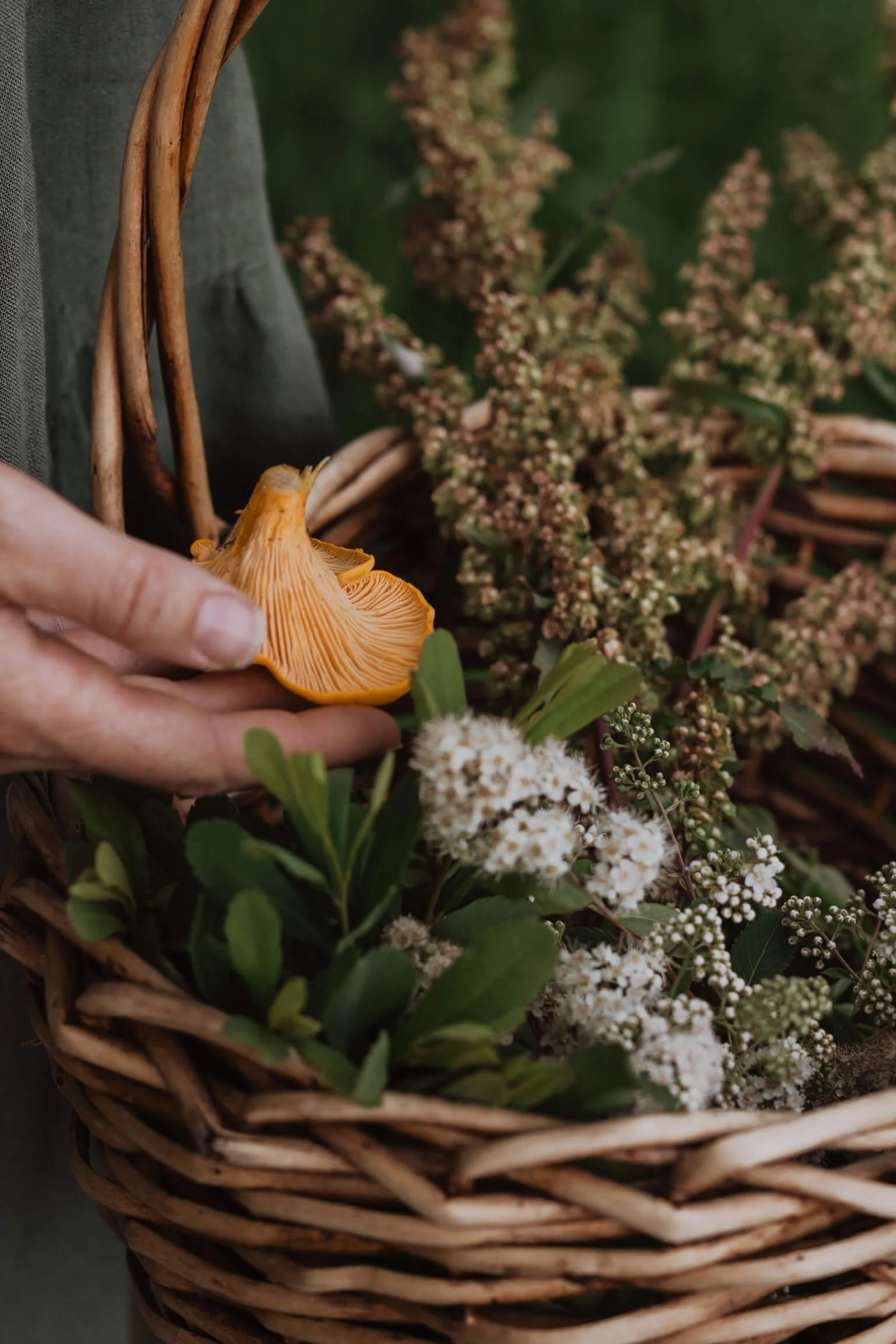Person holding an orange mushroom in a wicker basket filled with greenery and flowers.