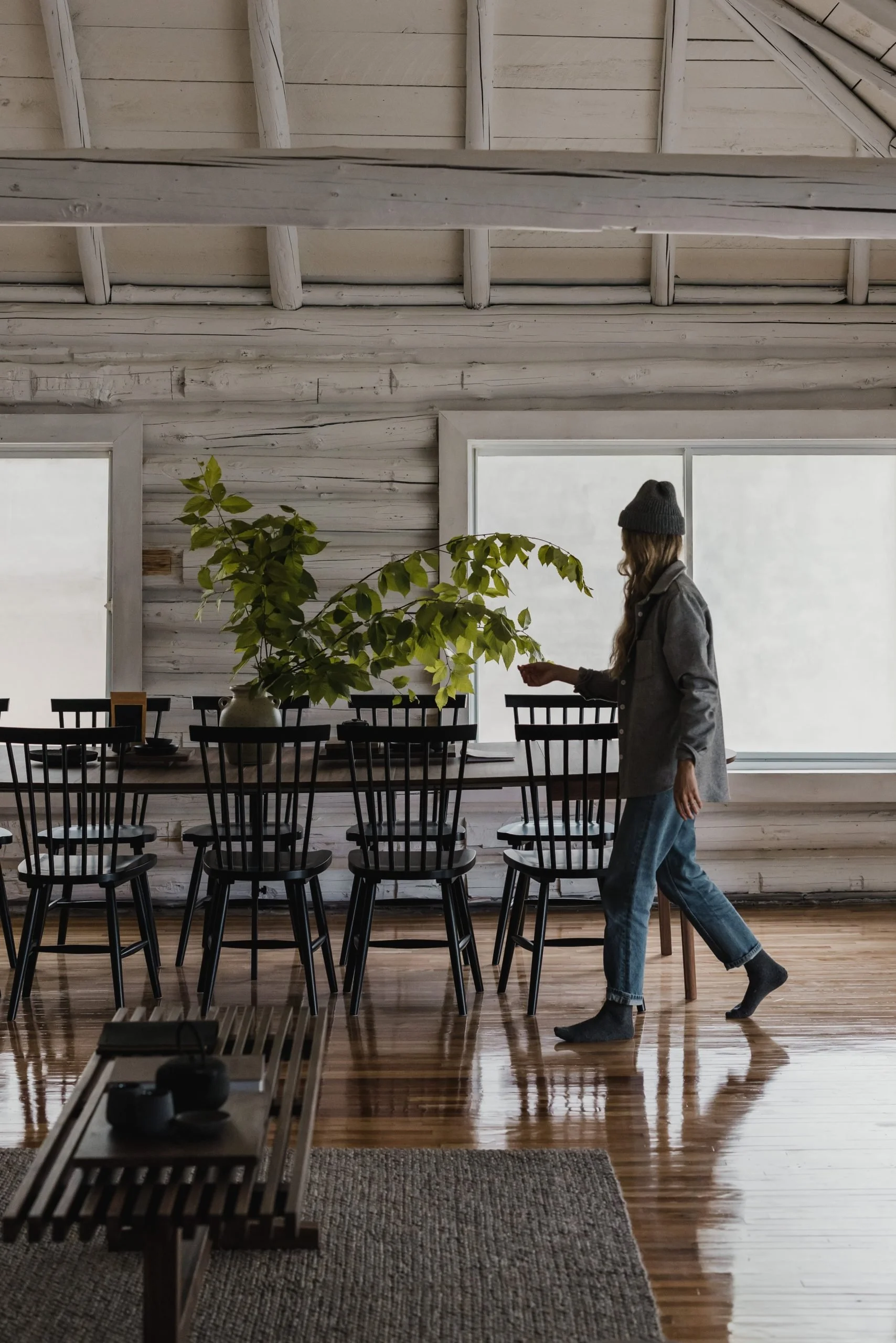 A woman in a gray coat, beanie, and jeans walks by a dining table inside a rustic-style room with wooden walls and floors. The table has black chairs and a large vase with green leafy branches.