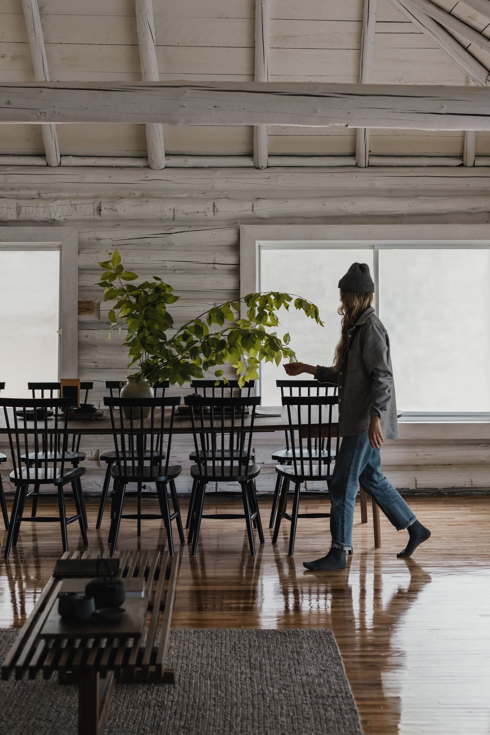 A woman in a gray coat, beanie, and jeans walks by a dining table inside a rustic-style room with wooden walls and floors. The table has black chairs and a large vase with green leafy branches.