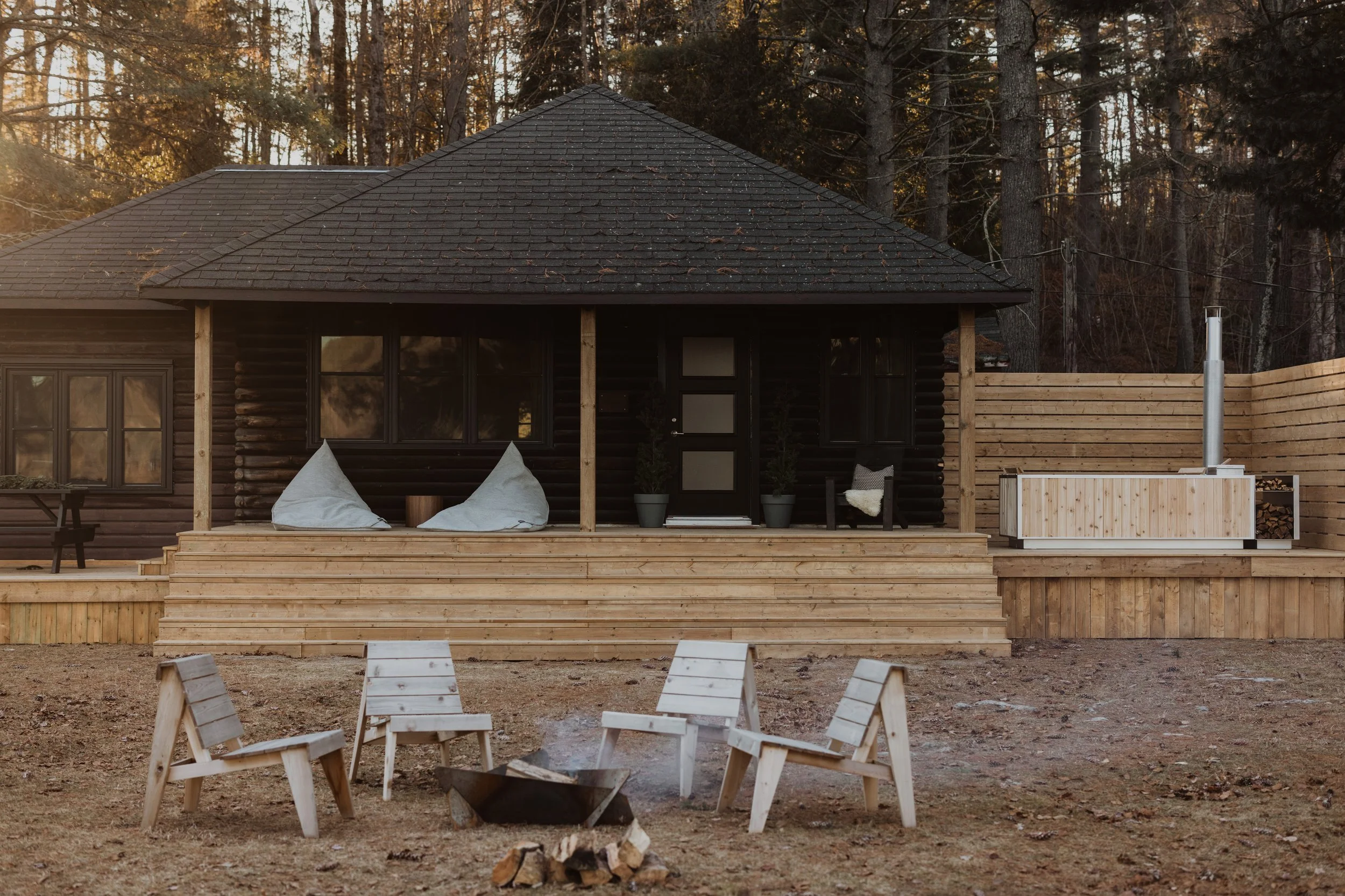 A wooden house with a deck and outdoor seating area in a forested setting during sunset, featuring three white chairs around a fire pit.