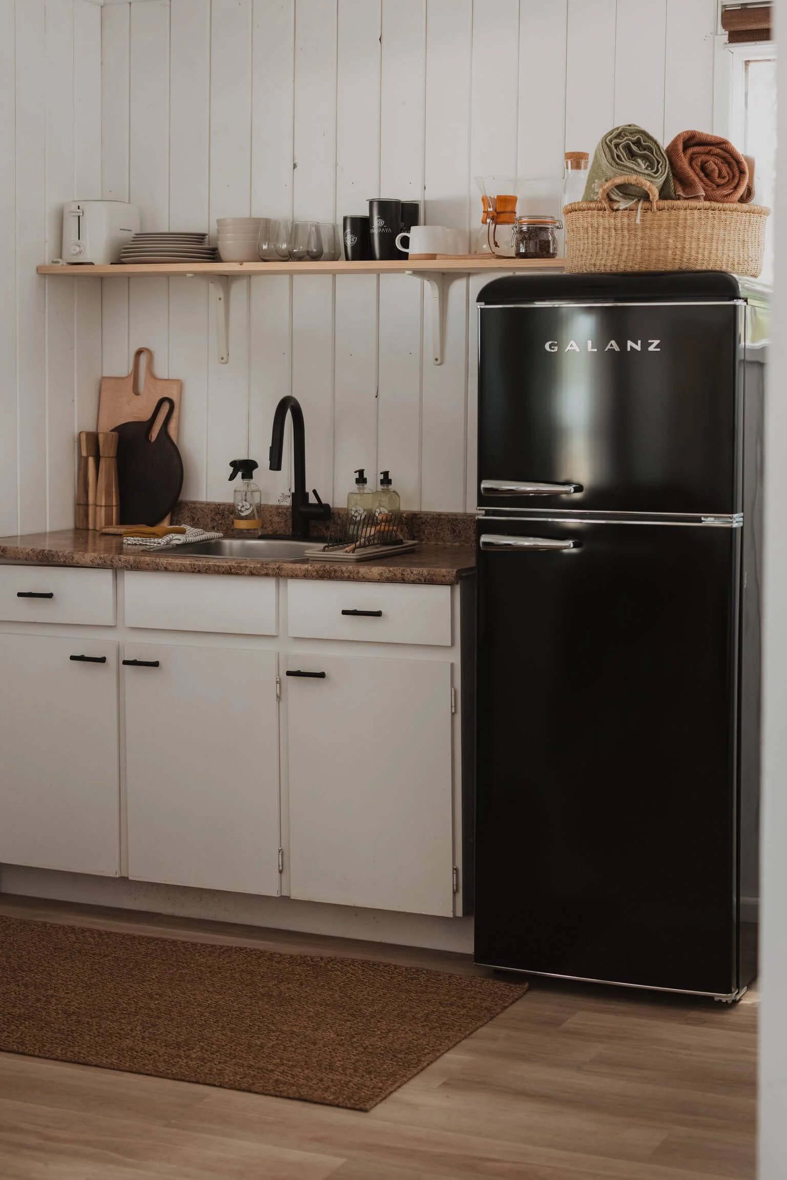 A kitchen corner with white cabinets, a black refrigerator, a brown countertop, a black faucet, and a wooden shelf holding dishes, glasses, and kitchen accessories.
