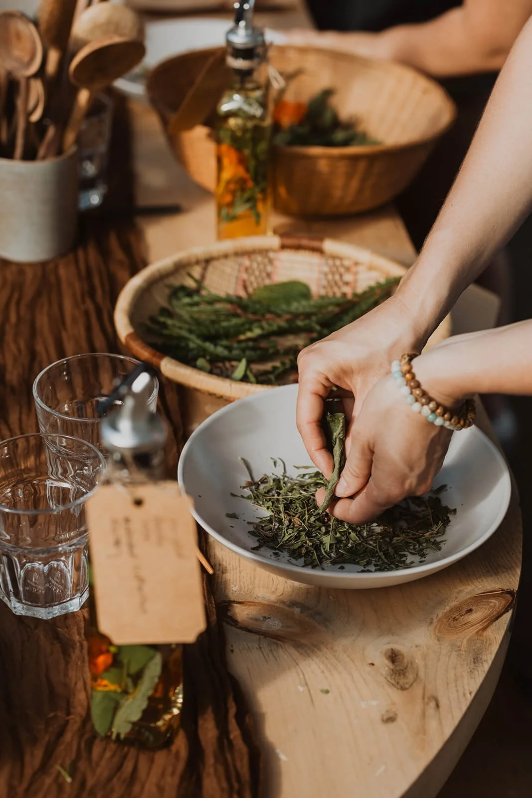 Person’s hands adding fresh herbs to a white bowl on a wooden table, with various herbs, glasses, and a bottle nearby.