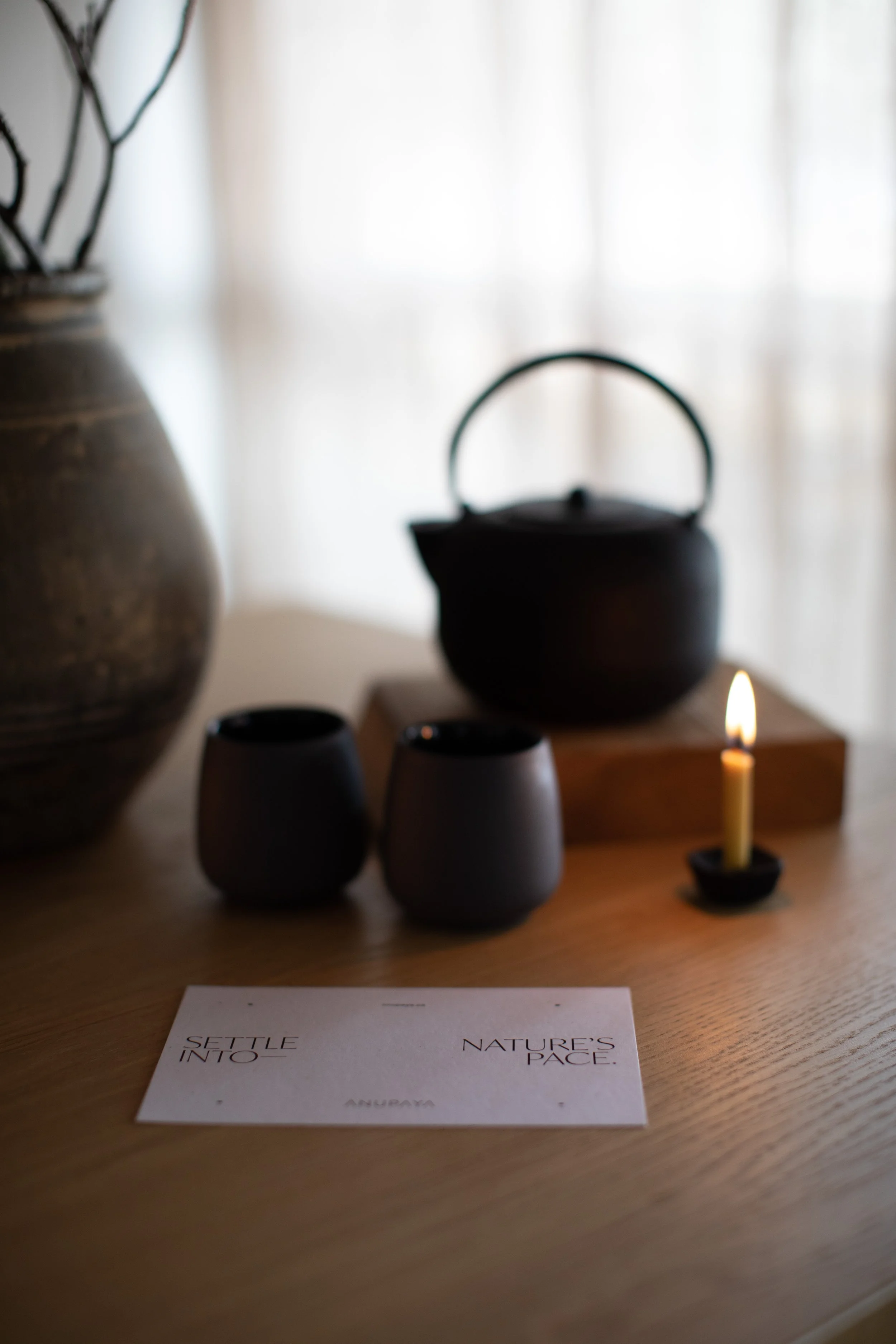 A table with a large brown vase, a black tea kettle on a wooden block, two black cups, and a small lit candle. A note card with the words 'Settle into Nature's Pace' and the name 'Anuraya.'