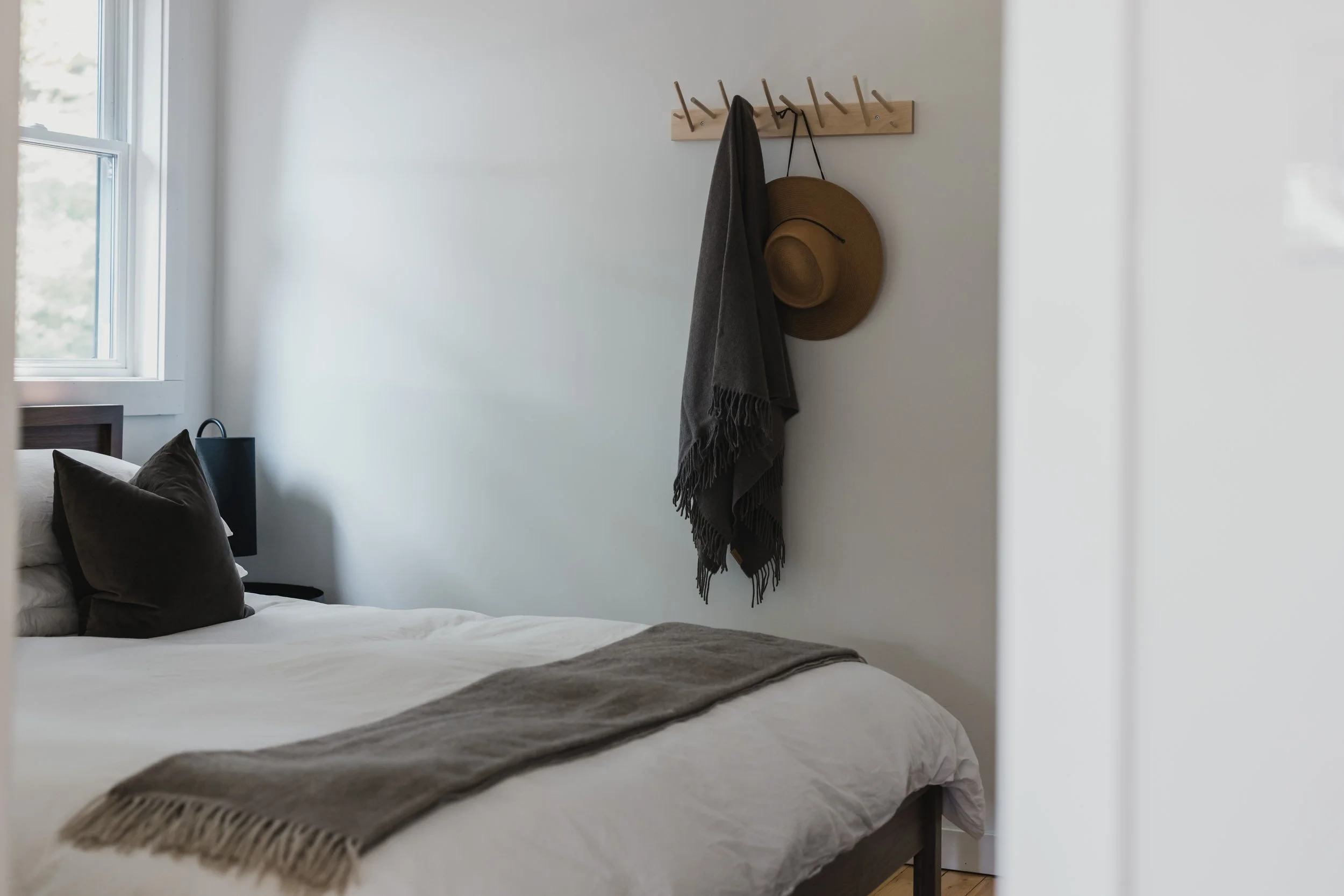 A cozy bedroom with a white bed along the wall, dark pillows, and a beige throw blanket. There is a window on the left and a wooden coat rack on the right wall holding a hat, a scarf, and a draped dark garment.