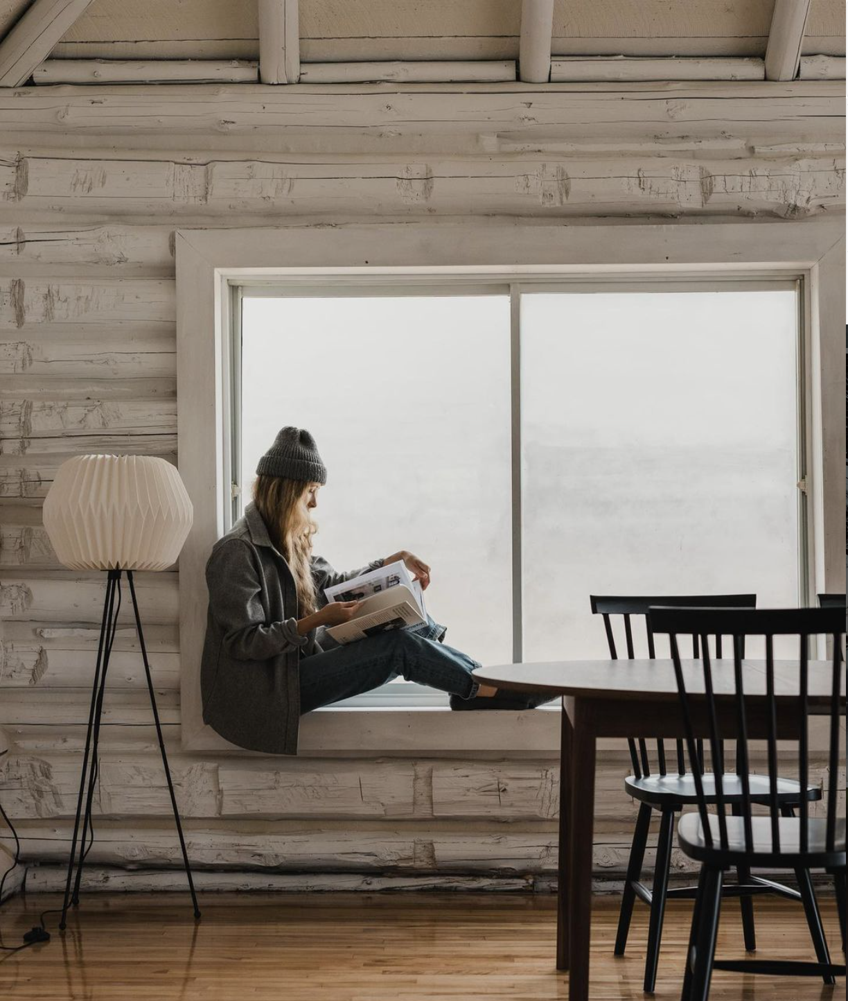 A young woman reading a book while sitting by a large window in a cozy, rustic room with white wooden walls and a wooden floor. She is wearing a gray coat, a gray beanie, and blue jeans. There is a tall, white lamp with a pleated shade to her left and a wooden dining table with black chairs to her right.