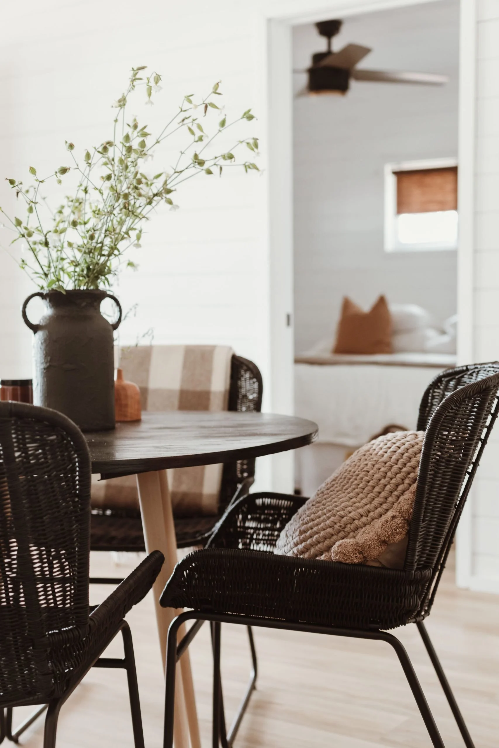 Dining room with a round wooden table, black wicker chairs, beige textured pillows, a large dark vase with greenery, and a cozy bedroom visible through a doorway in the background.