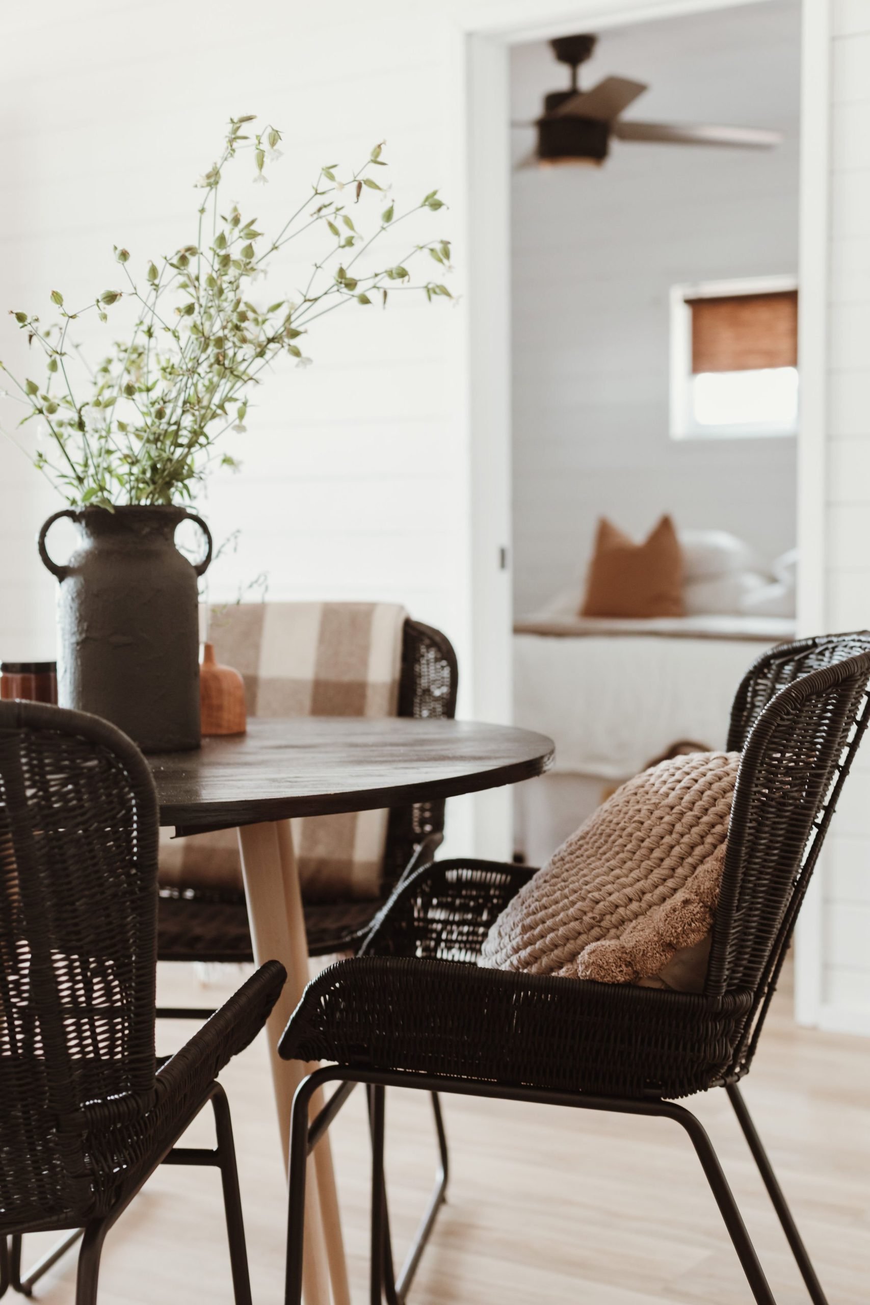 Dining room with a round wooden table, black wicker chairs, beige textured pillows, a large dark vase with greenery, and a cozy bedroom visible through a doorway in the background.