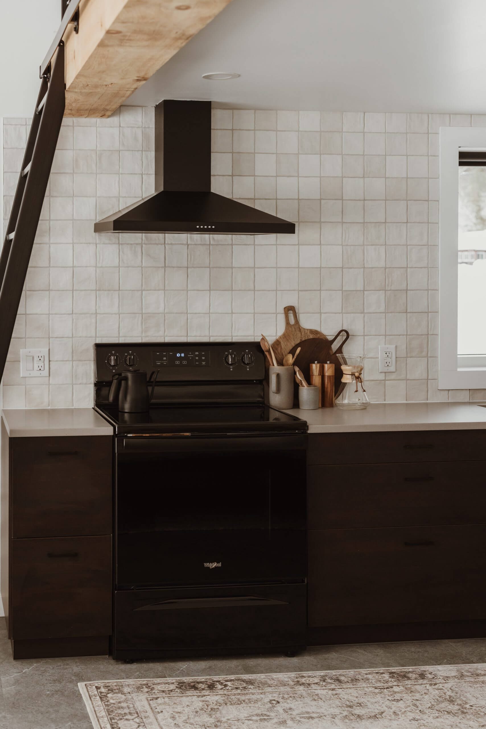 A modern kitchen with a black stove, a black range hood, and dark cabinetry. Decor includes wooden cutting boards, a gray pitcher, and various small kitchen items on the countertop. There is a window to the right and a patterned rug on the floor.