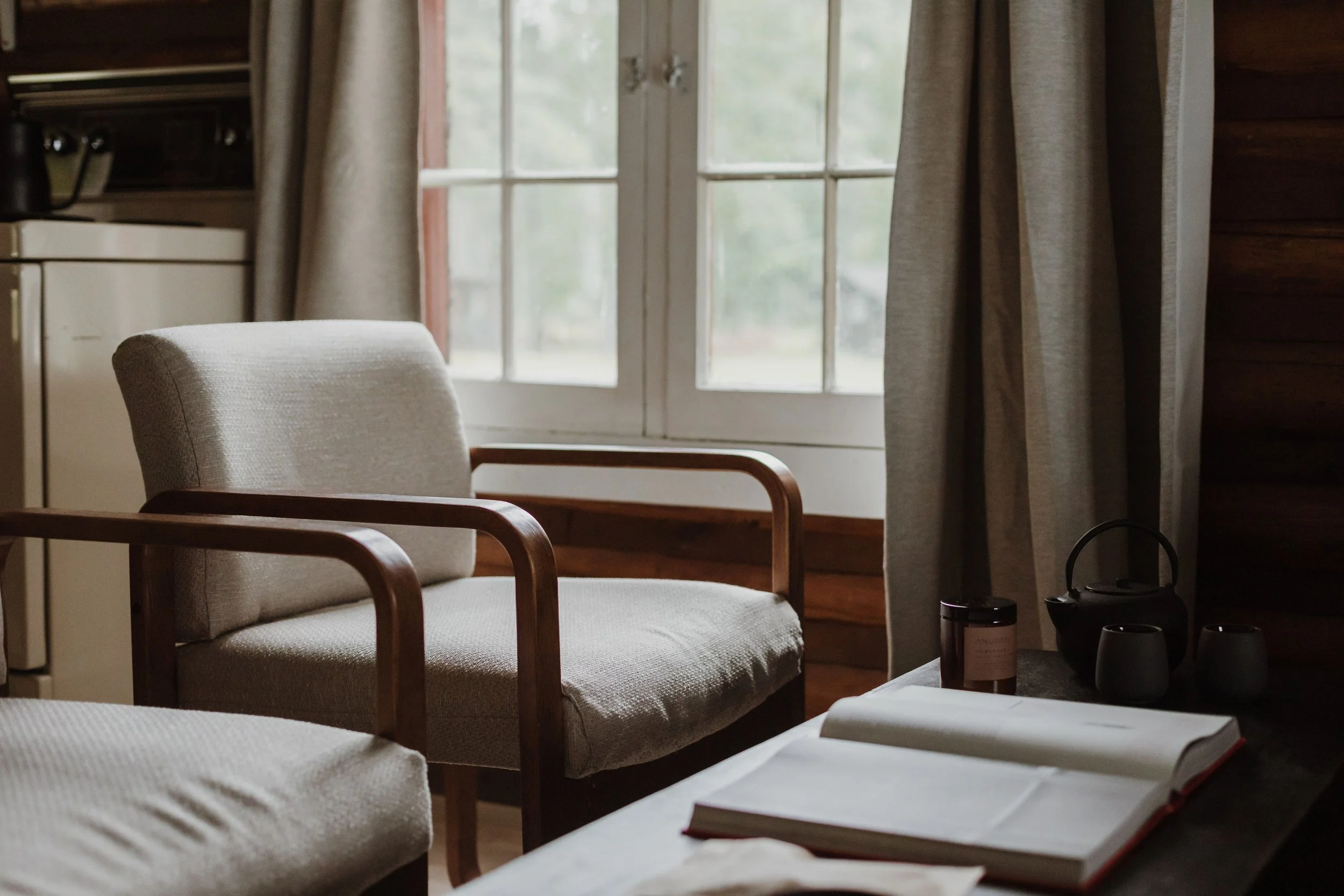 Cozy indoor space with a cushioned armchair near a window, a table with an open book, a teapot, cups, and a jar, and beige curtains.
