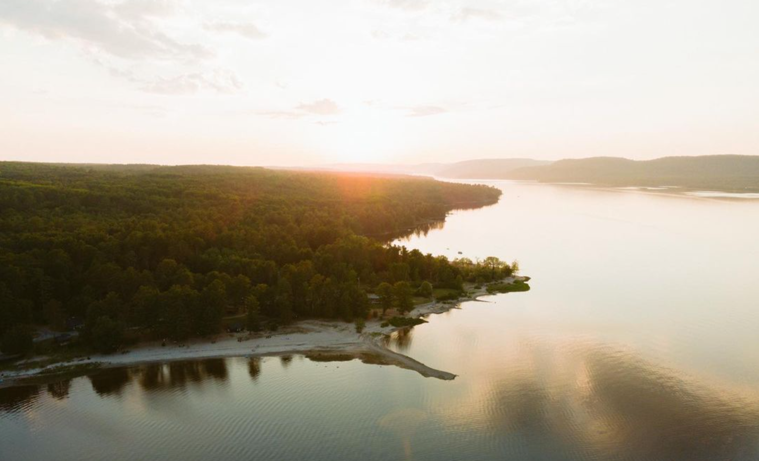 Aerial view of a river flowing through a forested landscape during sunset, with calm water reflecting the sky and distant hills.