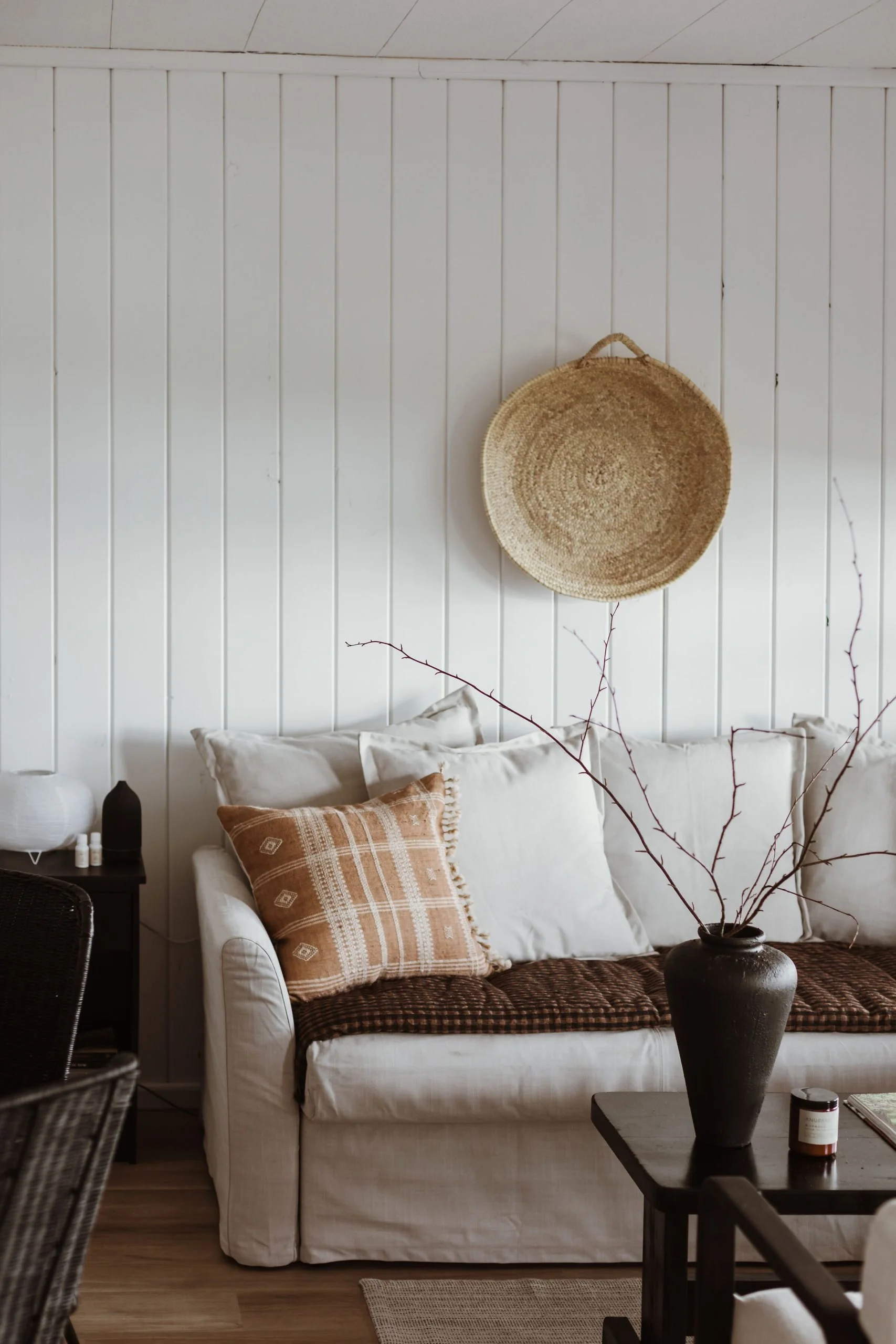 Cozy living room with white paneled wall, beige sofa with white and orange patterned pillows, black side table with decorative items, and a black vase with bare branches on a dark wooden coffee table.