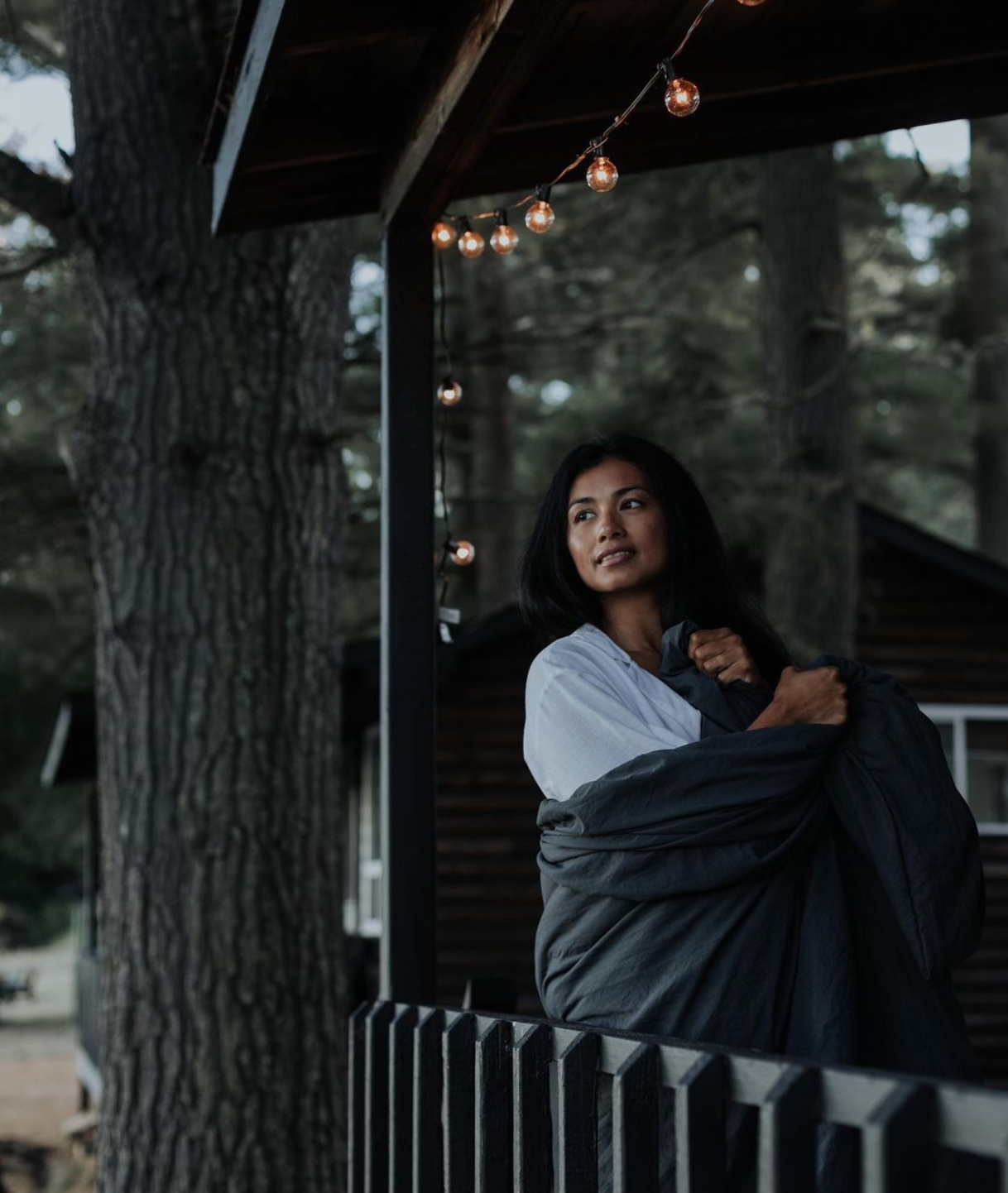 A woman standing on a porch with string lights, holding a black jacket, looking thoughtfully into the distance with trees in the background.