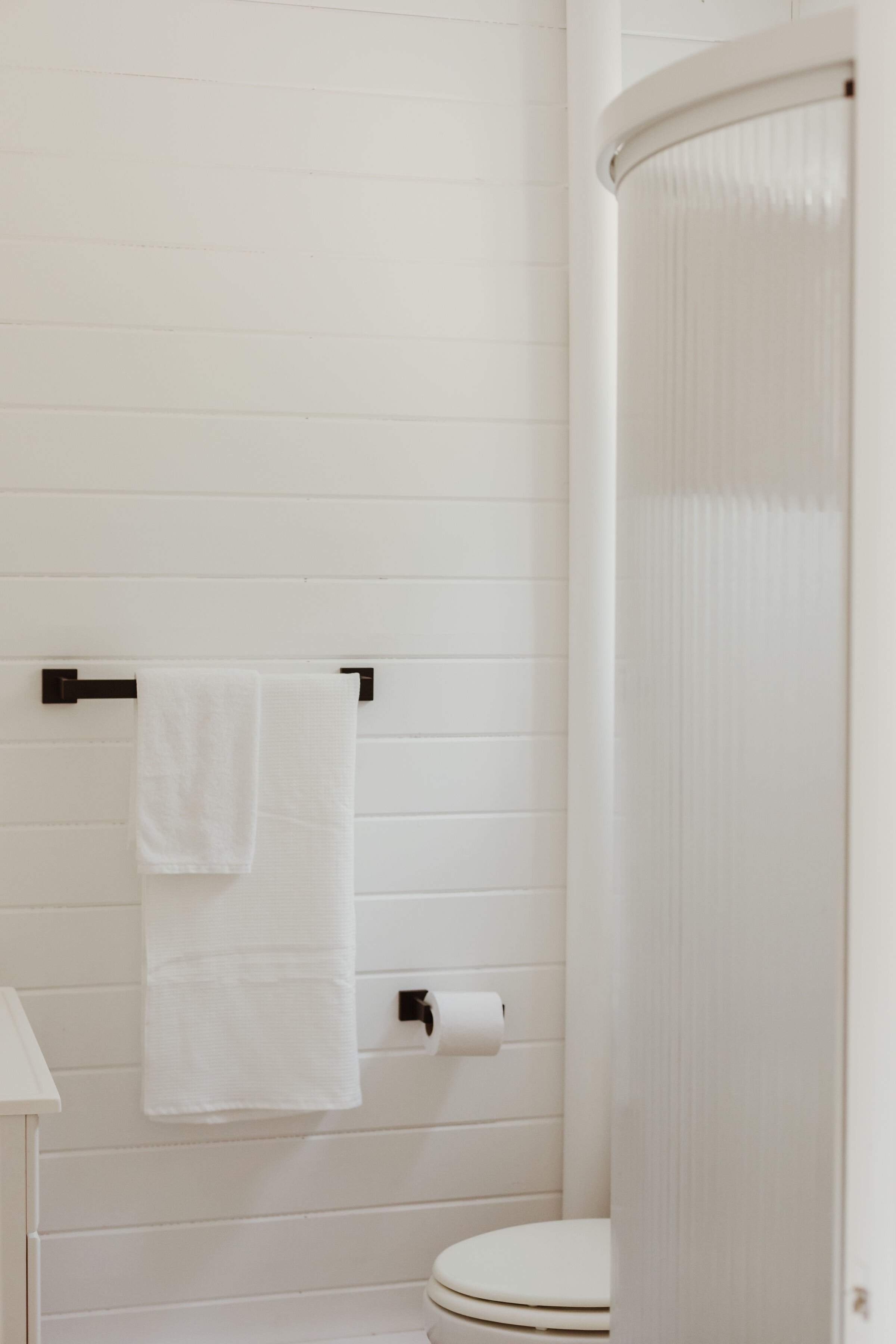 A minimalist bathroom with white wooden paneled walls, a white toilet, a black towel bar with a white towel, a toilet paper holder with a roll, and a curved shower stall with a ribbed white door.