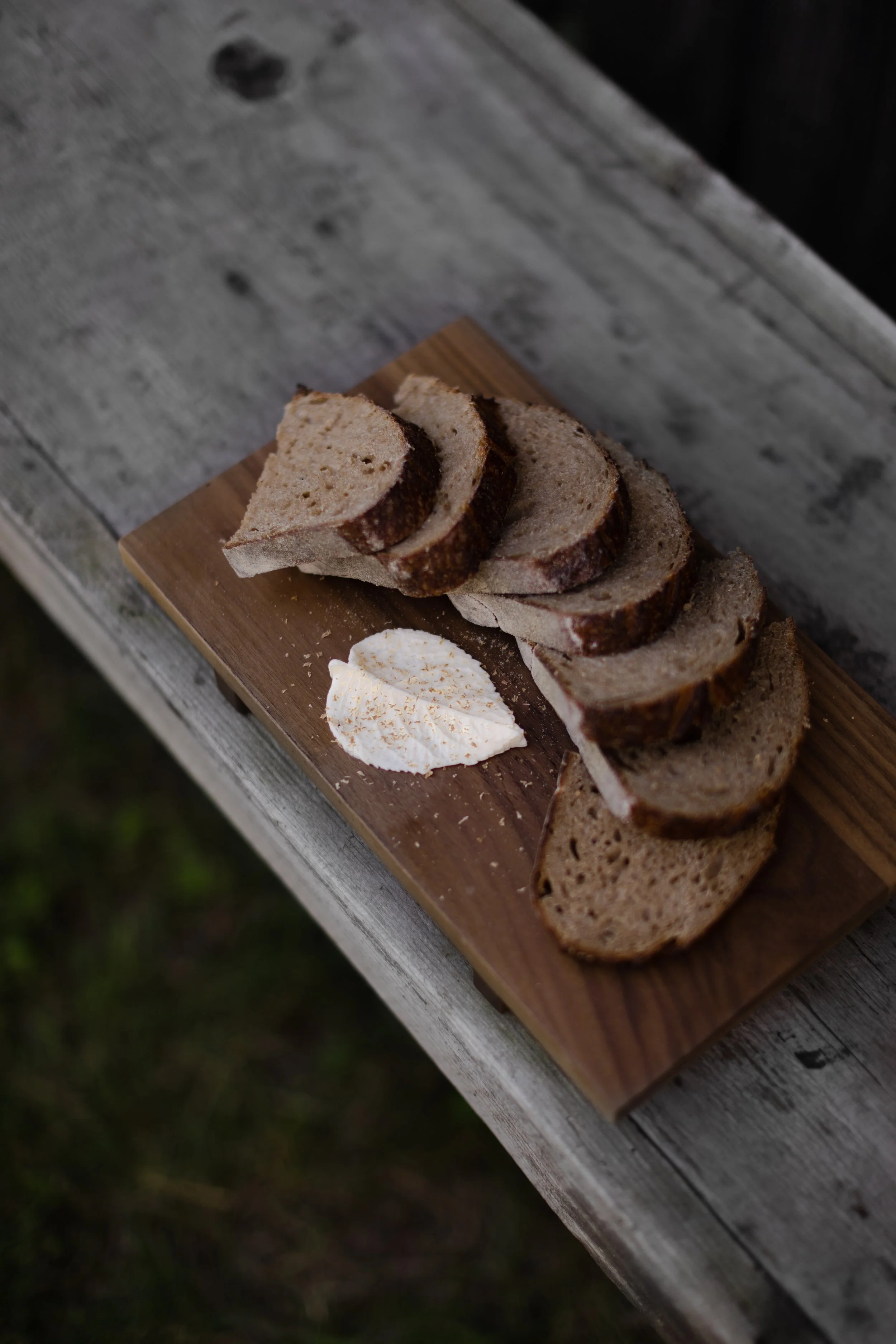 Slices of rustic bread on a wooden cutting board with a dollop of cream cheese or butter garnished with a small sprinkle of spice, set on a weathered wooden table.