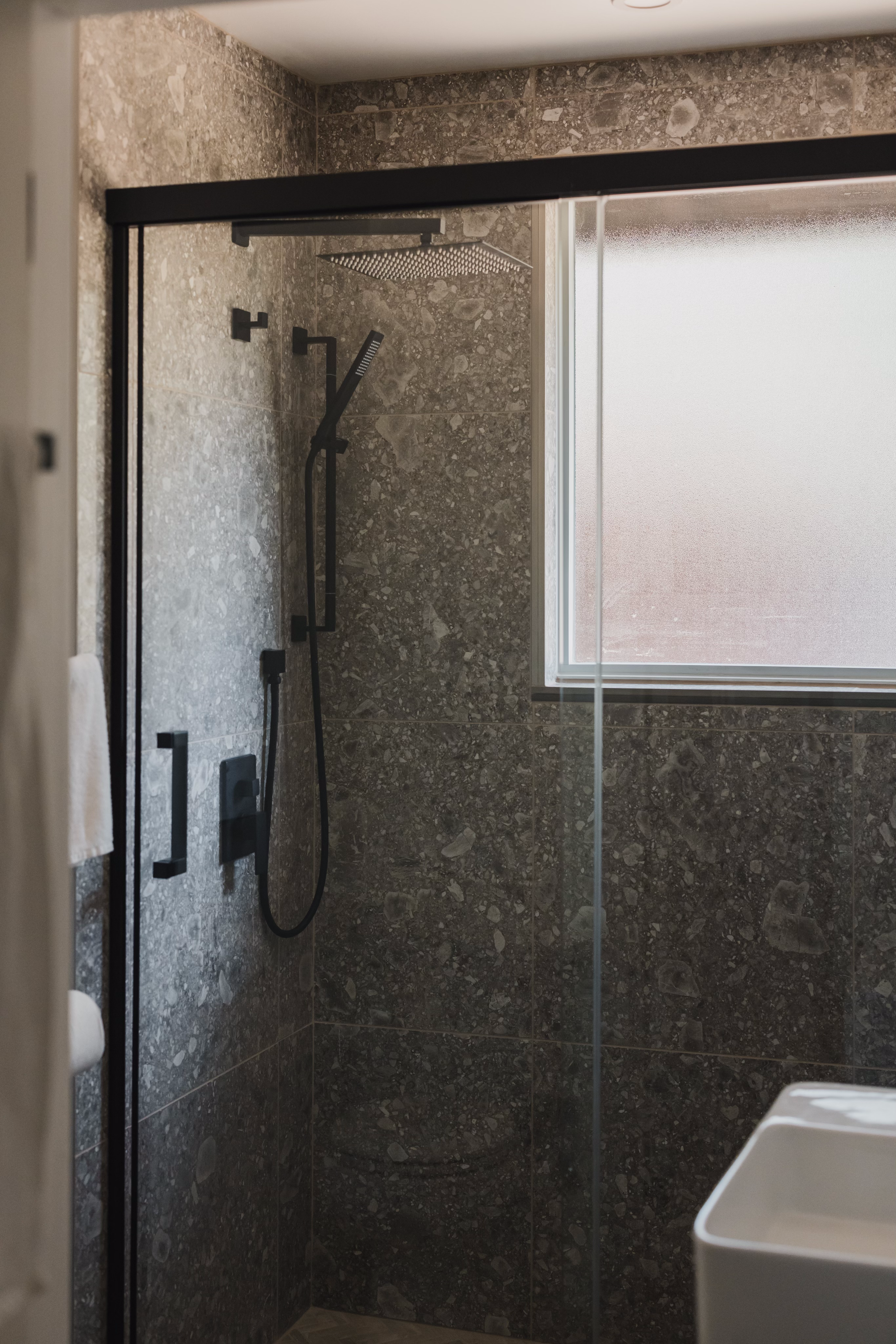 Modern bathroom with a glass enclosed shower featuring dark stone tiles, a black rain showerhead, a handheld shower, and a frosted window.
