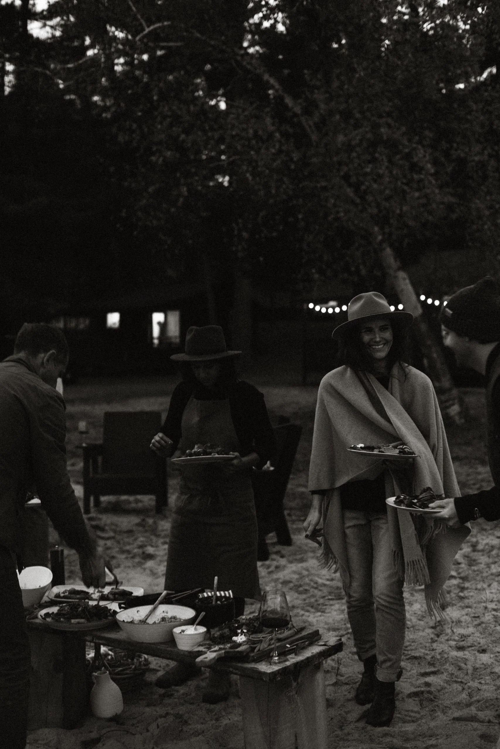 People having a dinner outdoors at night, with a table of food and drinks, some wearing hats, in a natural setting with trees and string lights.