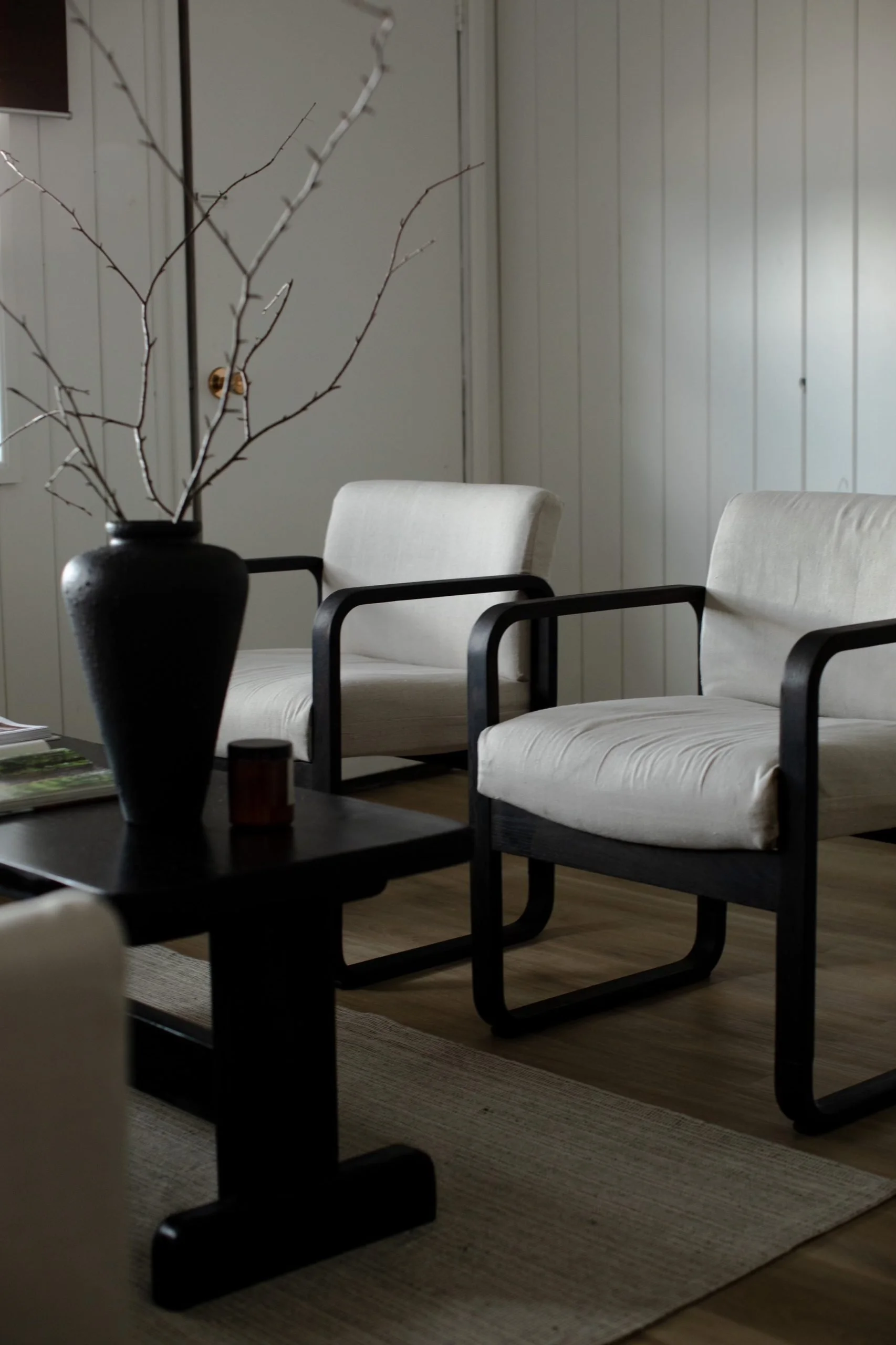 Living room with two white cushioned armchairs with black wooden frames, a black side table with a black vase containing dried branches, a small candle, and some magazines, on a beige rug with wooden flooring and white paneled walls.