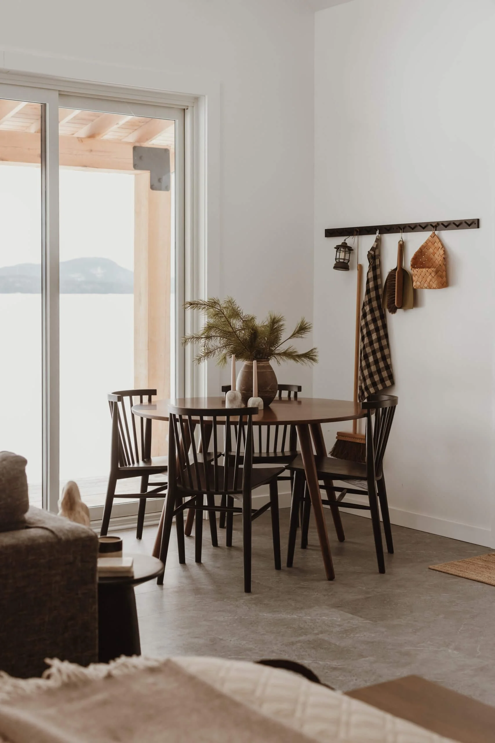 A cozy dining area with a round wooden table surrounded by black chairs, decorated with a fall-inspired centerpiece. The space features a large sliding glass door leading to a balcony with a view of water and distant land, and a wall with hooks holding various rustic decorative items and tools.