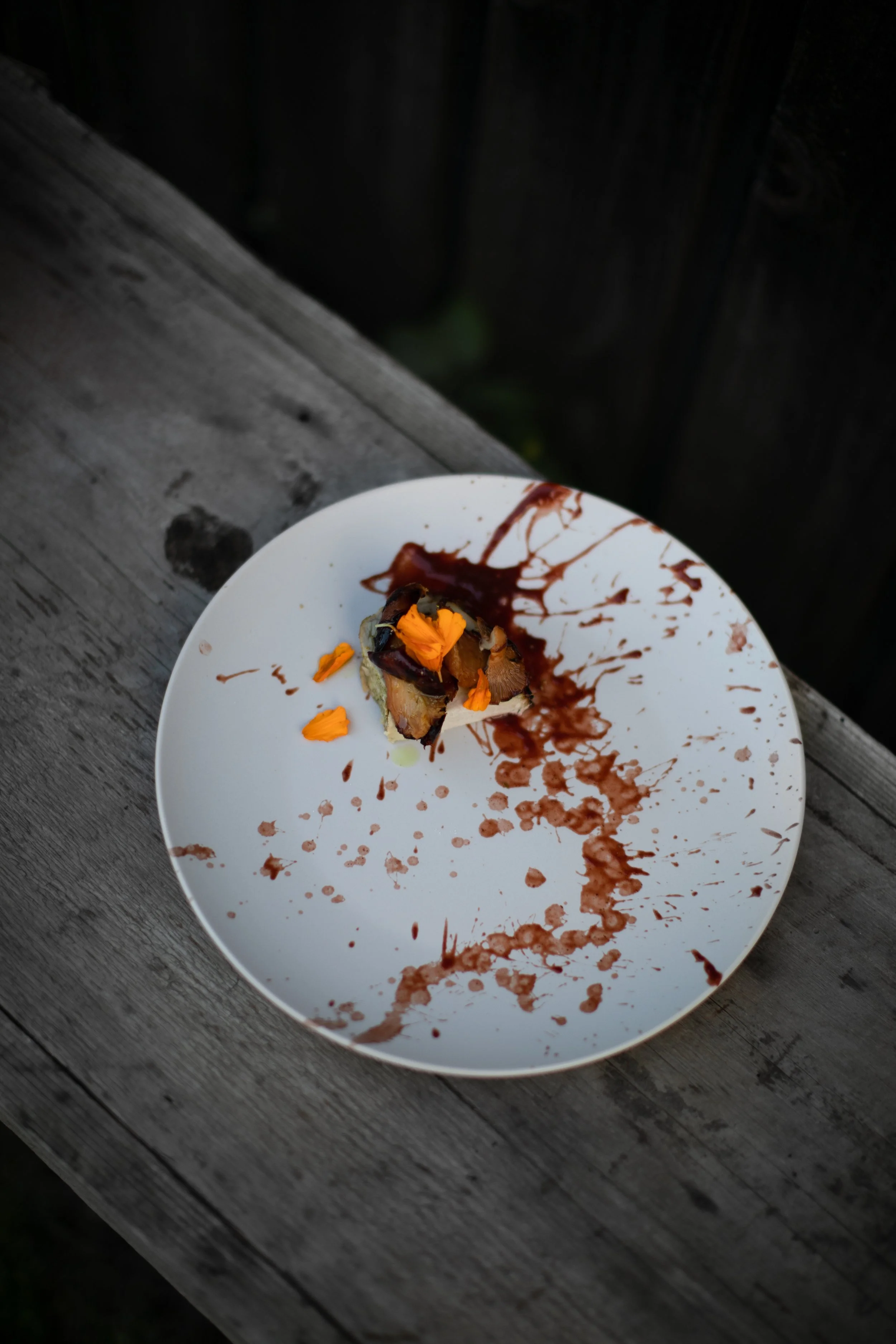 A white plate with a small portion of food, topped with orange flower petals, on a weathered wooden surface. Brown sauce or chocolate is splattered across the plate and wooden surface.