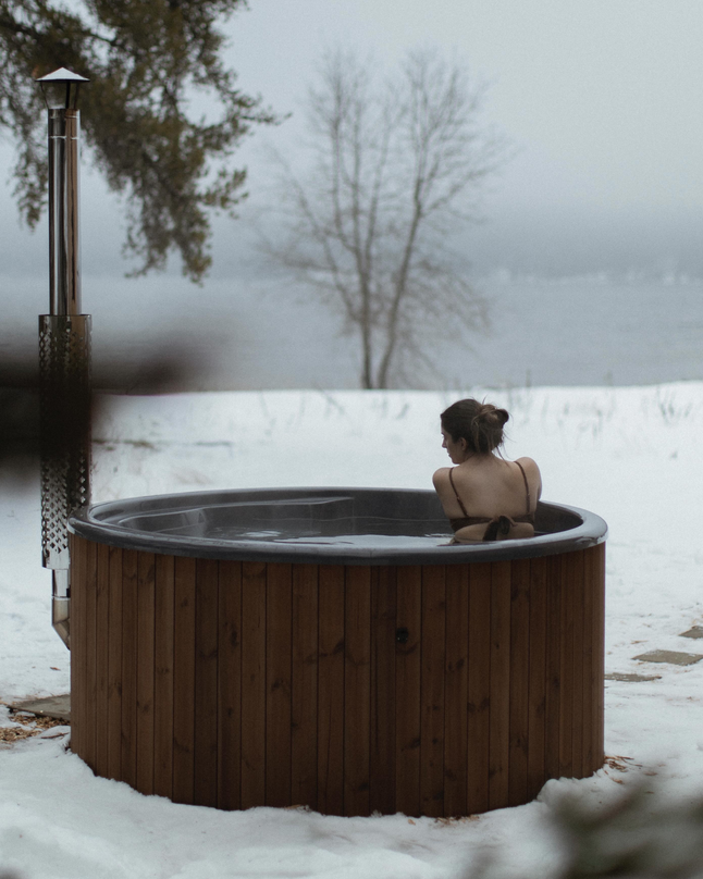 A woman with her hair in a bun soaking in a wooden hot tub outdoors on a snowy landscape with trees in the background and a foggy atmosphere