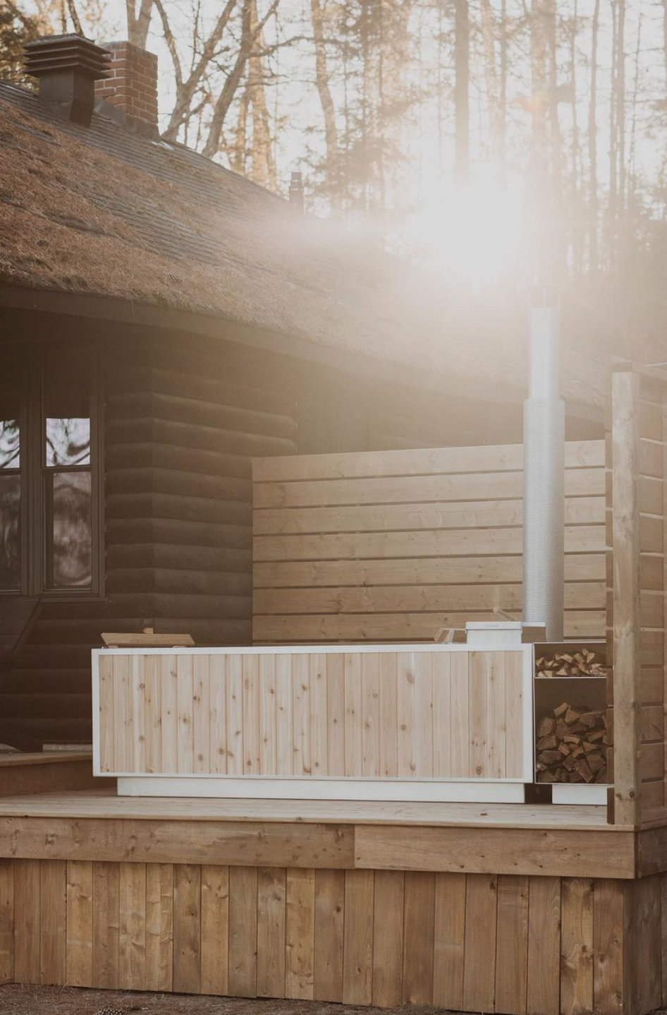 Wooden outdoor structure with a log cabin wall, a wood-fired hot tub, and a wood storage area, set against a backdrop of trees and sunlight.