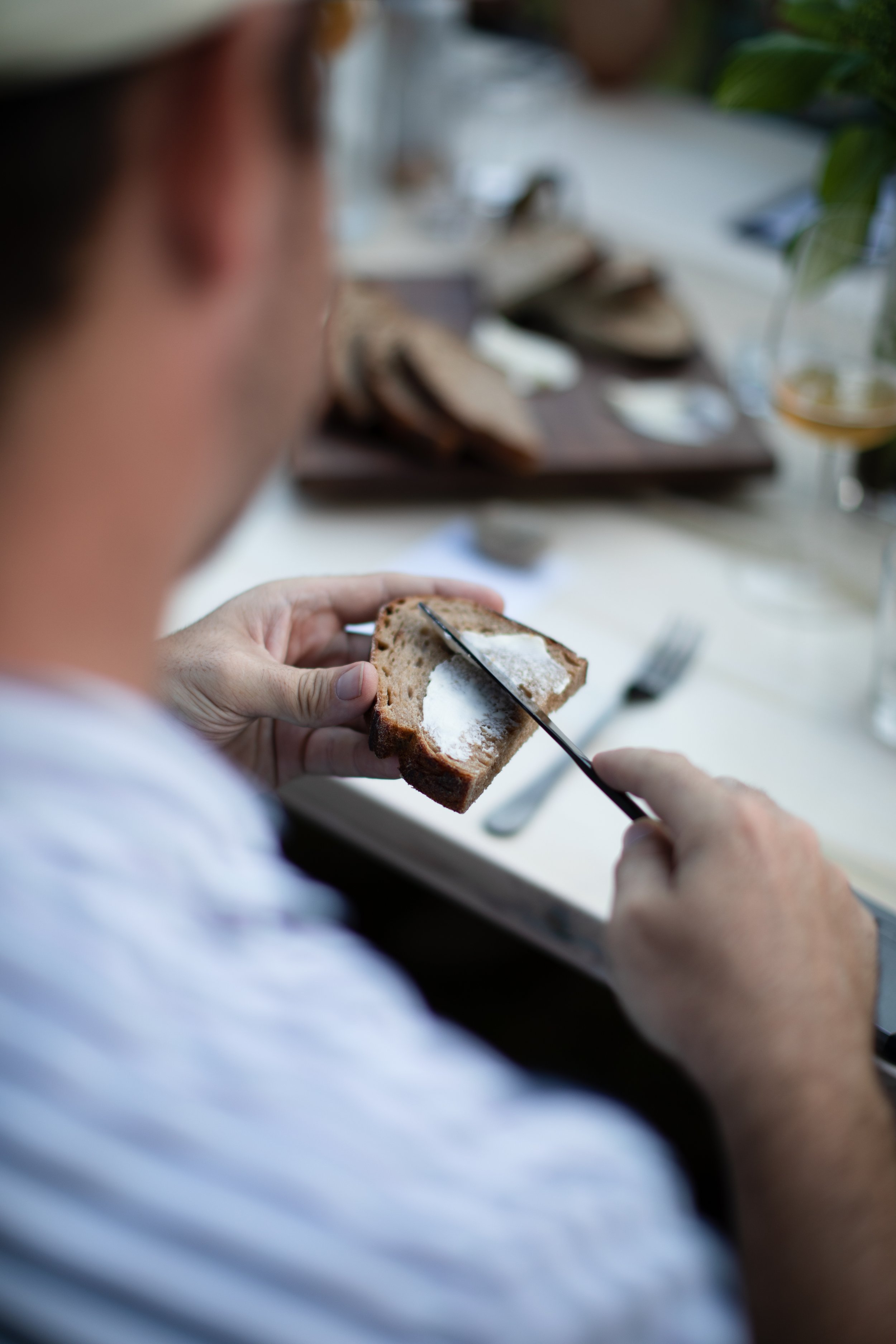 A person spreading salt on a slice of bread with a knife, sitting at a table with a blurred background of various food items and utensils.