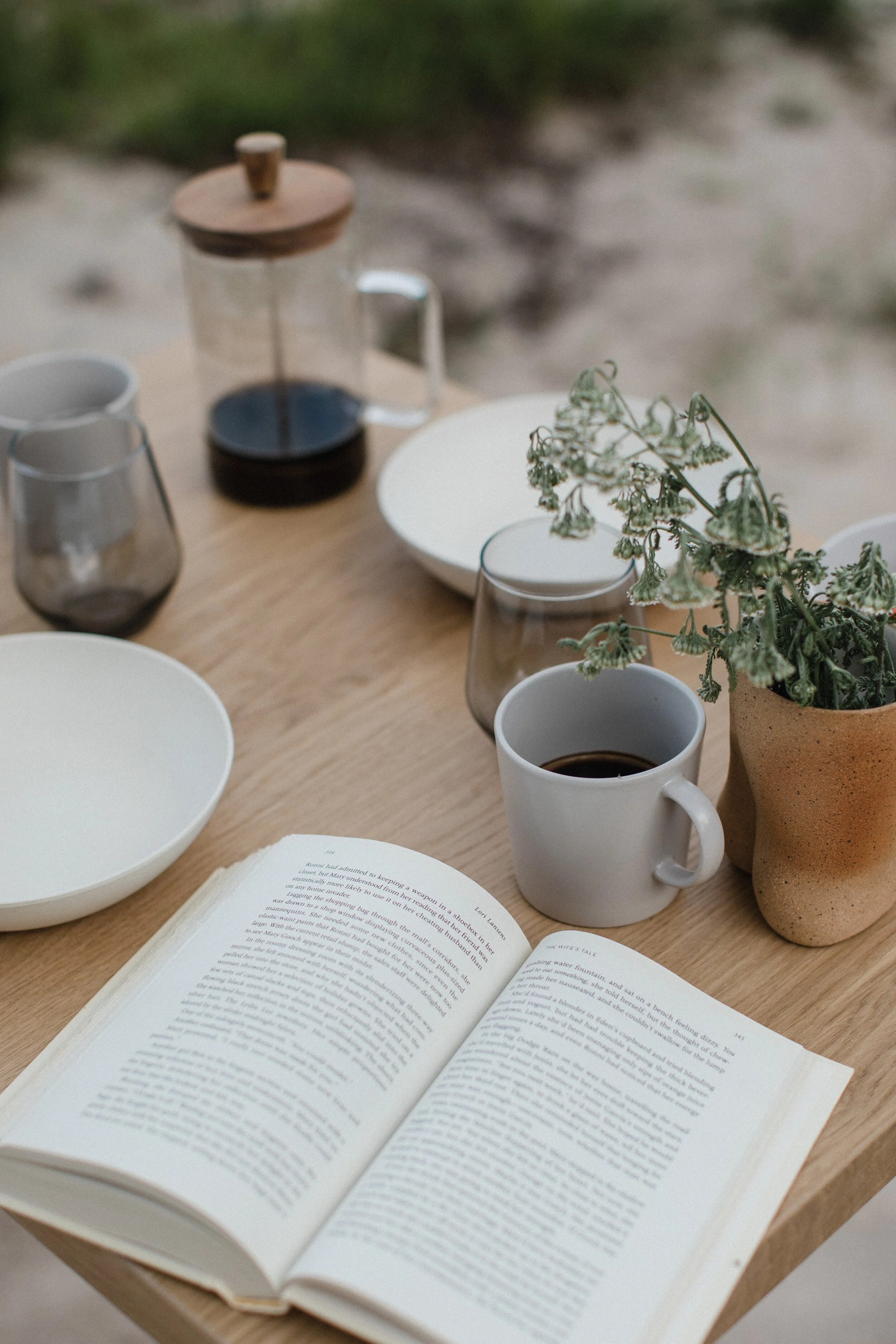 A wooden table set outdoors with a book, coffee mug filled with coffee, empty different glasses, a small pot with green plants, a white bowl, and a French press with coffee.