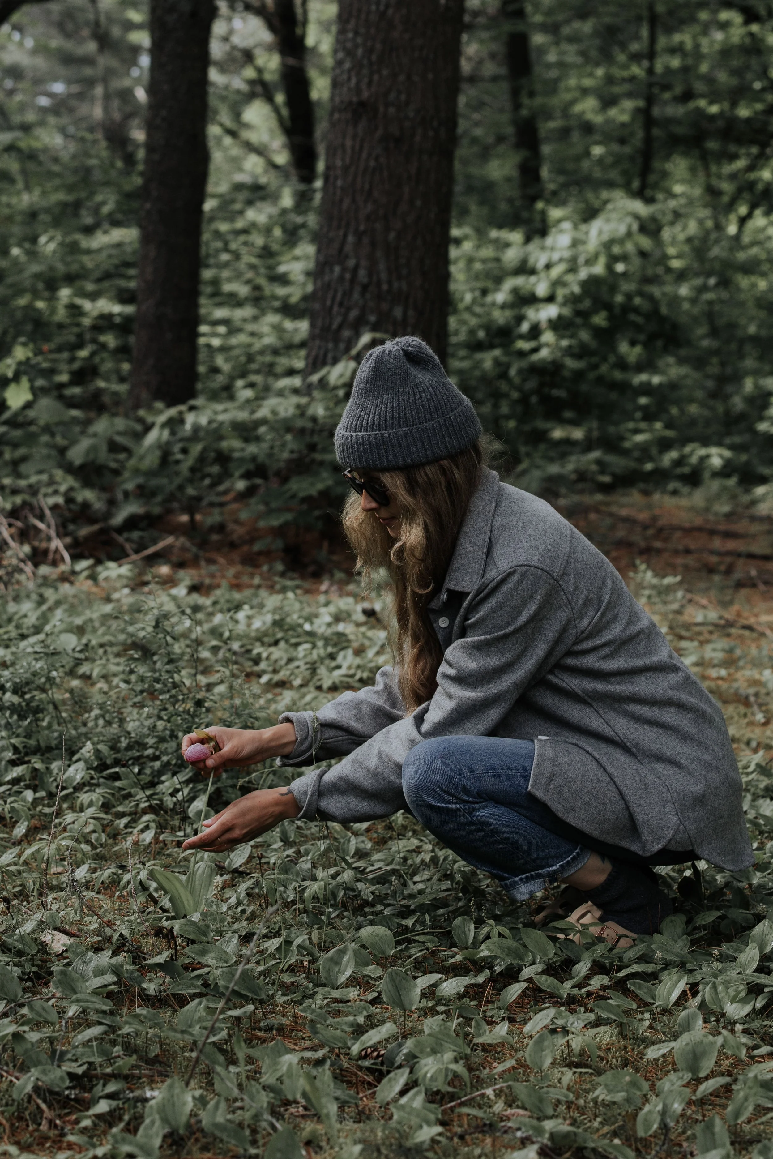 A woman in a gray coat, beanie, and sunglasses squatting in a forest, holding a flower in her hands.