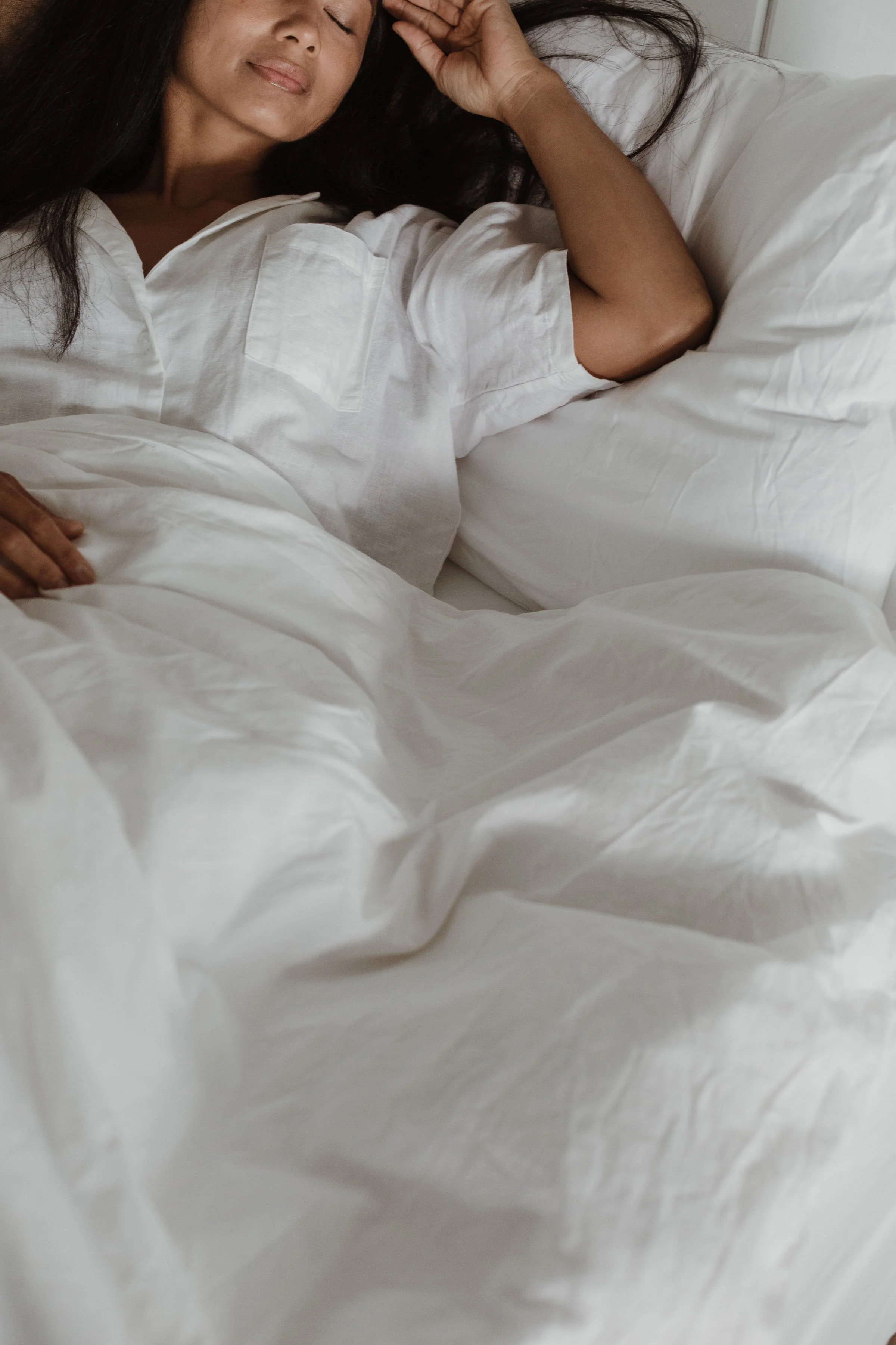 A woman with dark hair sleeping in a white bed, wearing a white shirt, with her eyes closed and a peaceful expression.