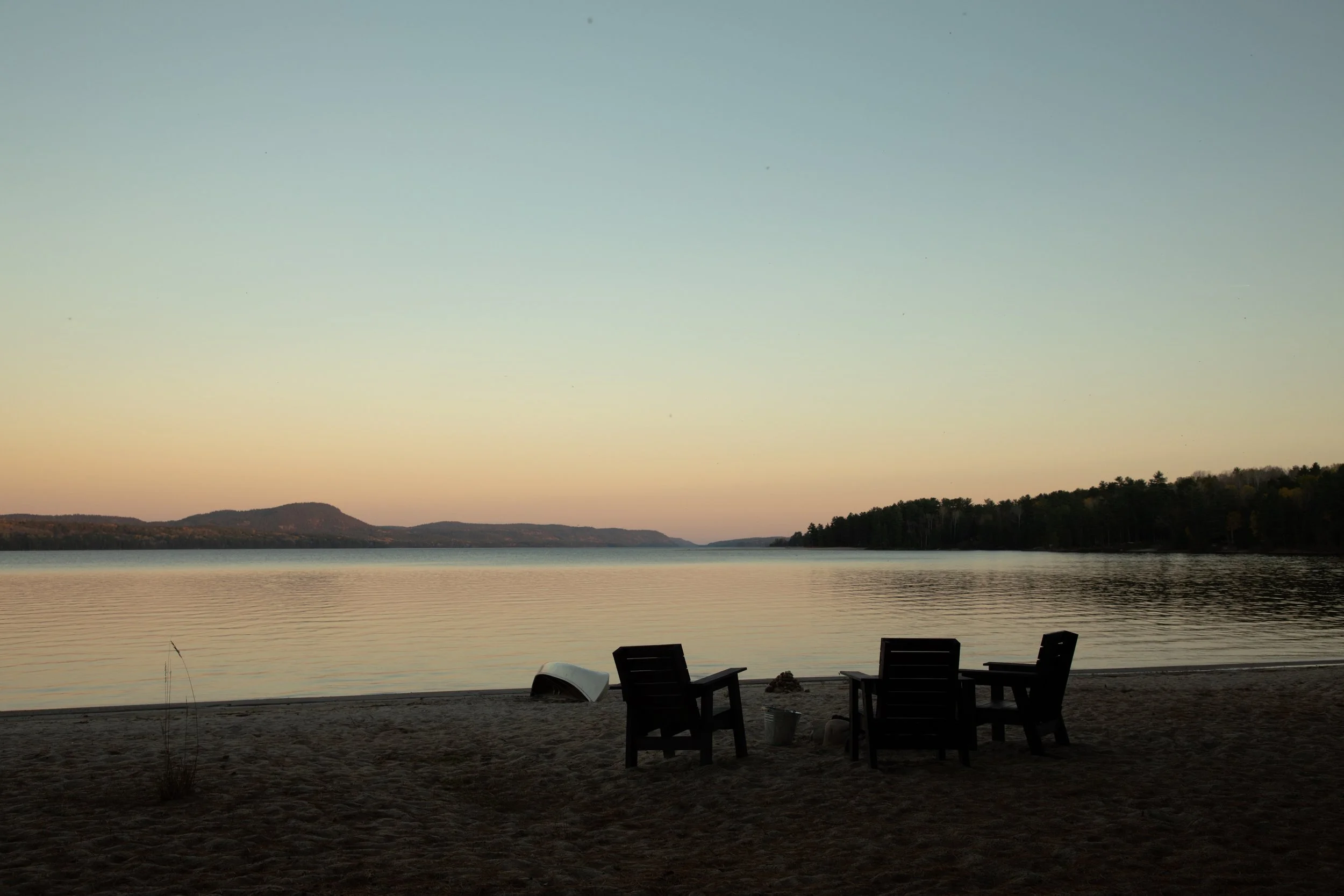 A peaceful lakeside scene at sunset with three empty chairs on the sandy beach, a small white boat overturned nearby, calm water, and distant hills with trees under a pastel sky.