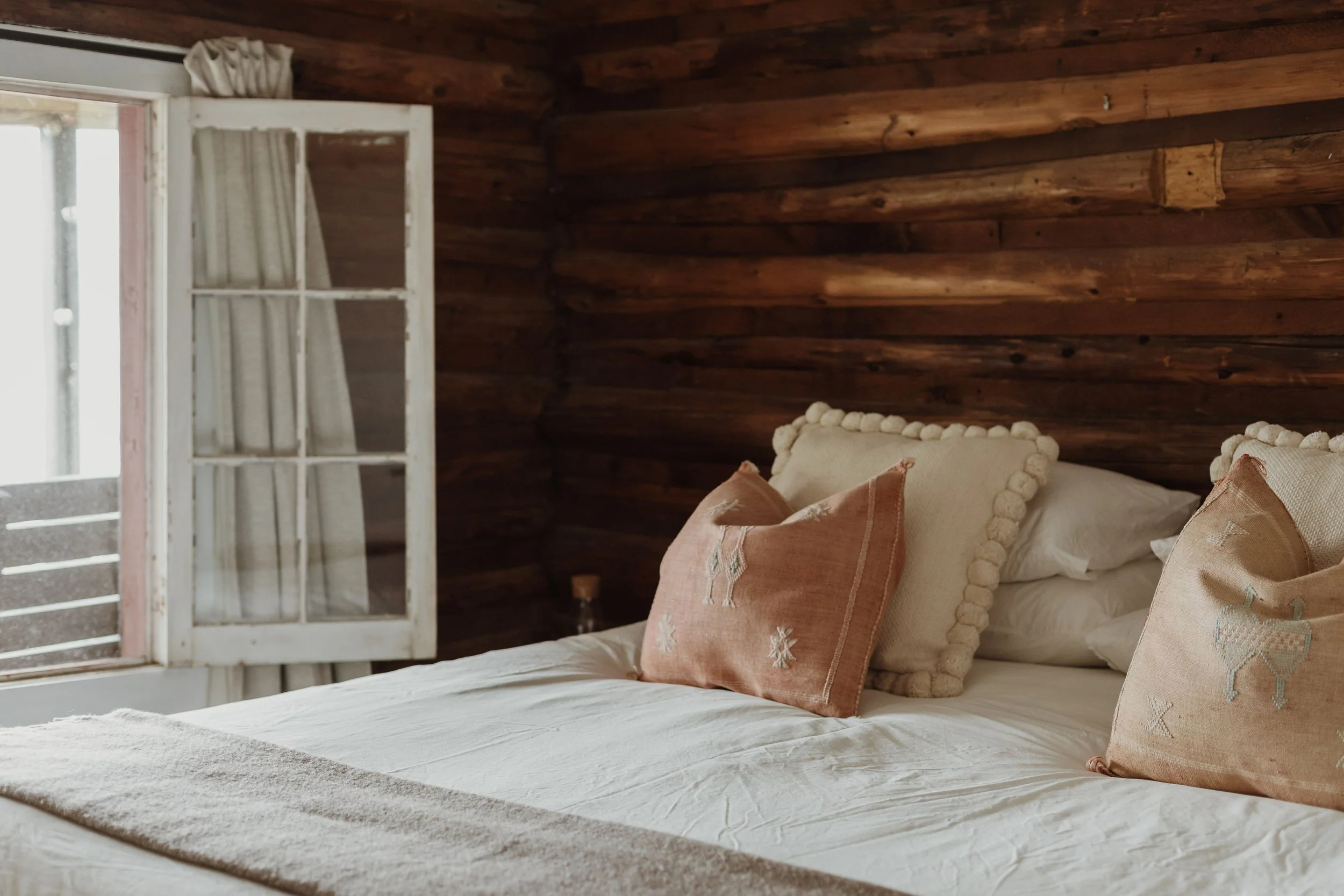 A cozy bedroom with a wooden wall, open window with white curtains, and a neatly made bed with embroidered pillows.