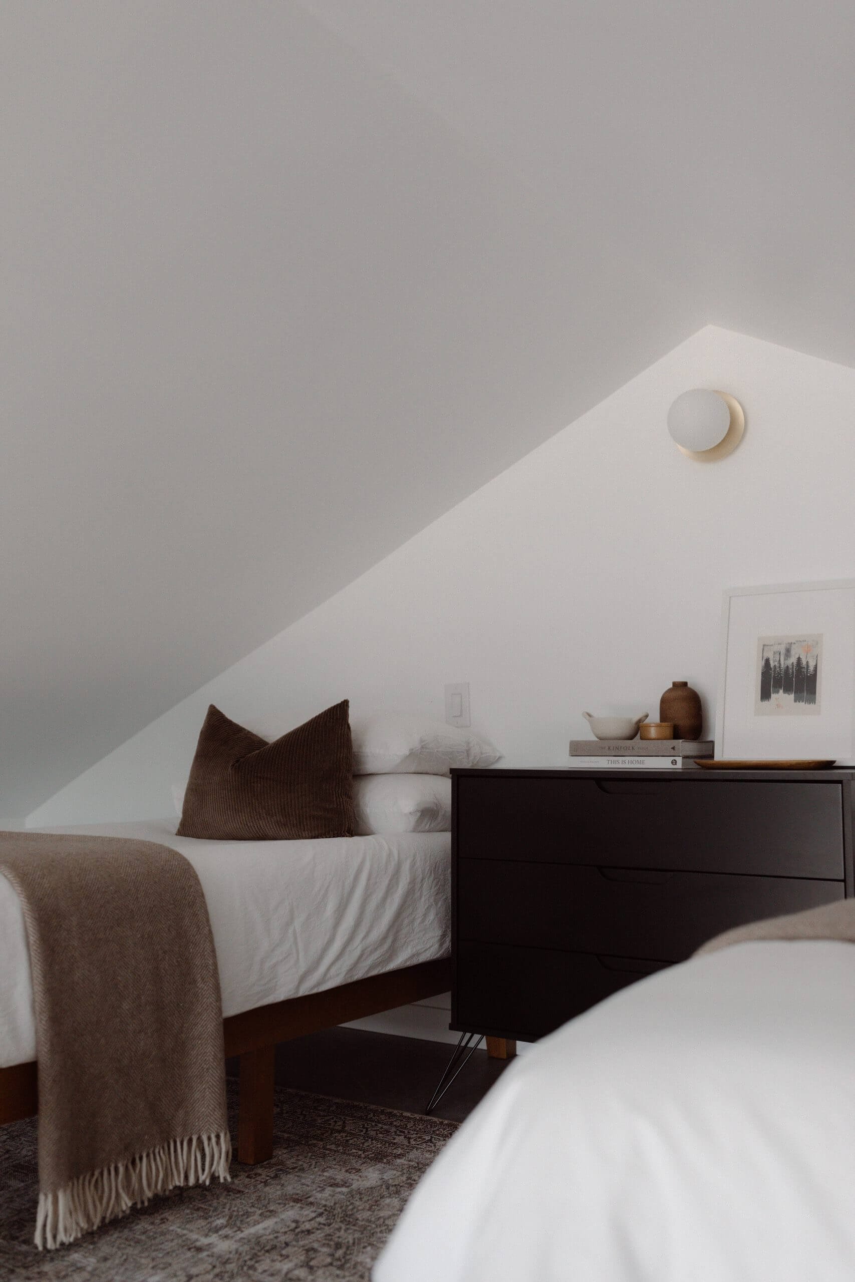 A cozy bedroom with sloped white ceiling, two beds with white bedding, and a black dresser decorated with books, a white bowl, and brown vases. One bed is partially visible in the foreground, while the other is against the wall with a brown pillow and blanket.