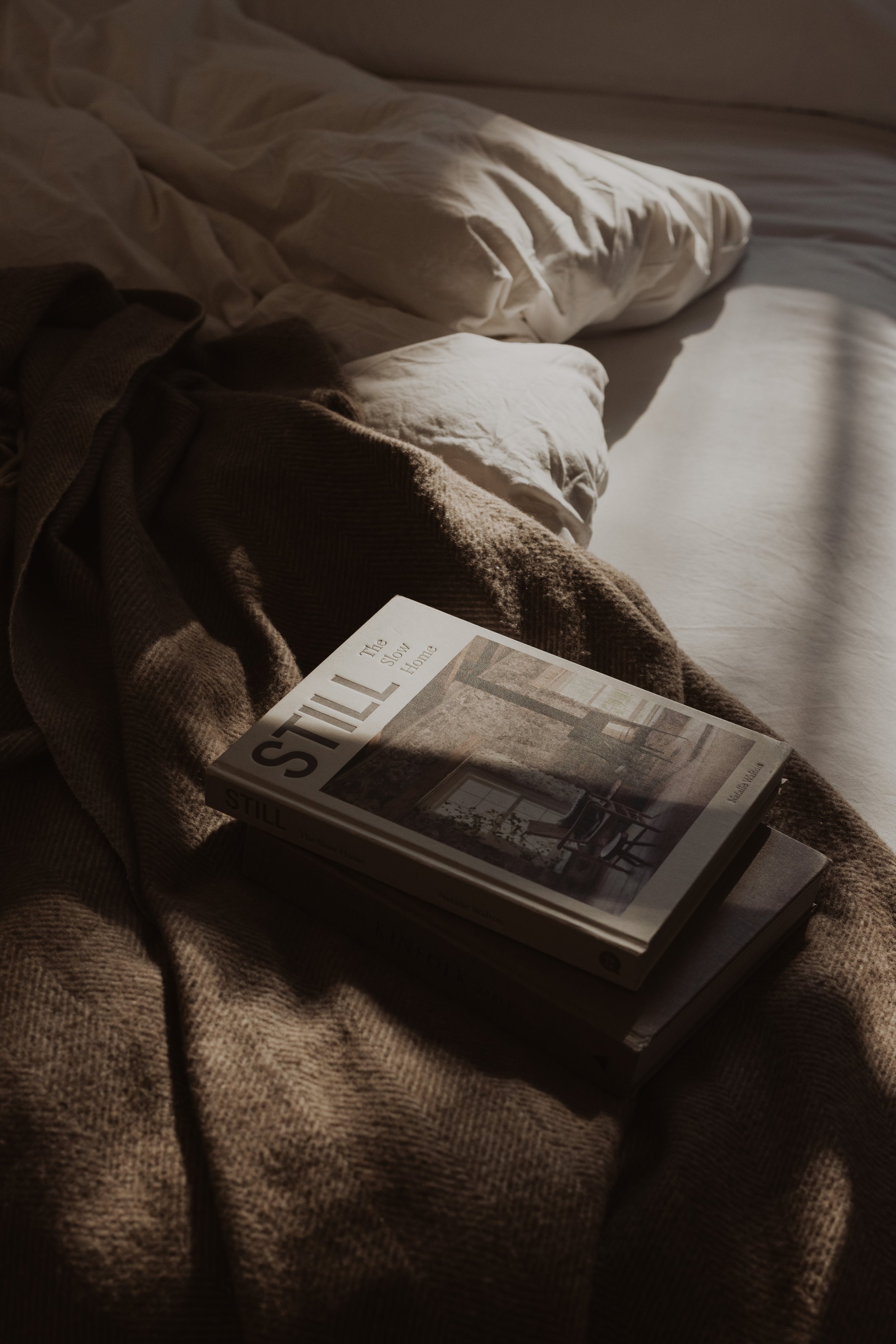 A cozy bed with white and beige linens, a brown blanket, and a book titled 'Still' resting on top, illuminated by soft, natural light.