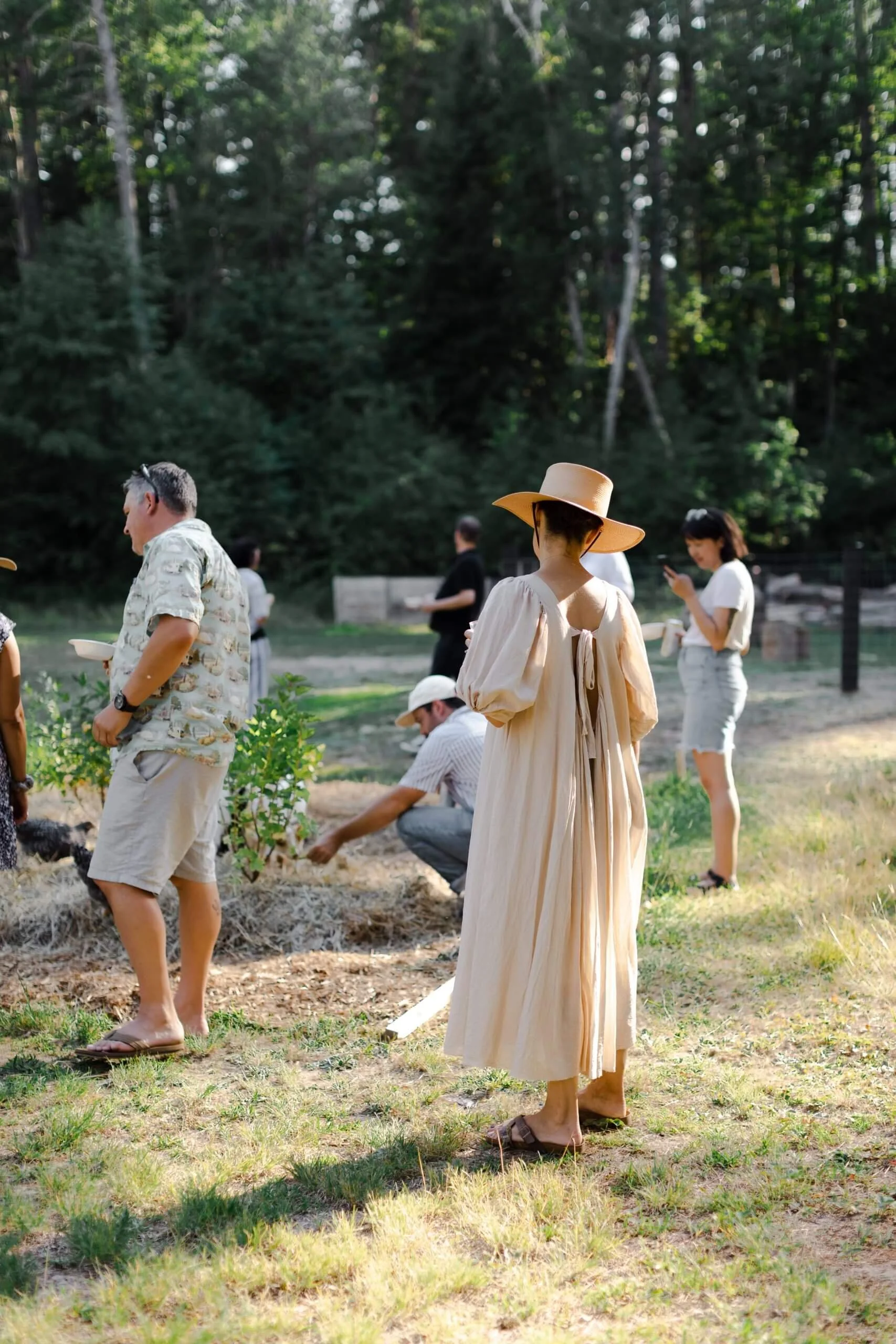 People gather outdoors in a grassy area with trees in the background. One woman in a flowing beige dress and wide-brimmed hat stands in the foreground, while others are engaged in gardening and socializing.