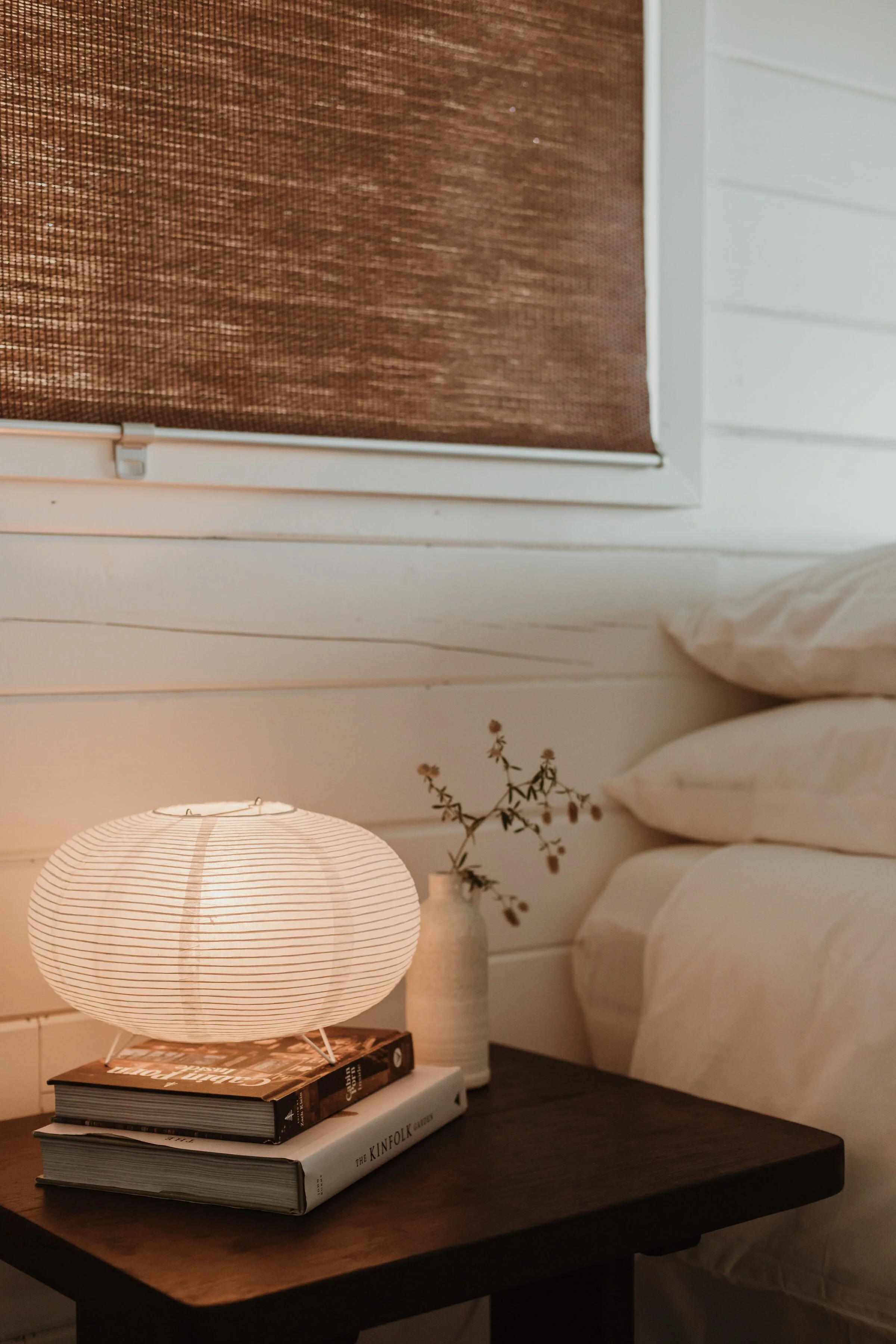 A cozy nightstand scene with a warm white paper lantern on top of two stacked books, a small white vase with dried wildflowers, and behind it a beige sofa and a neutral wall with a window covered by a brown woven shade.