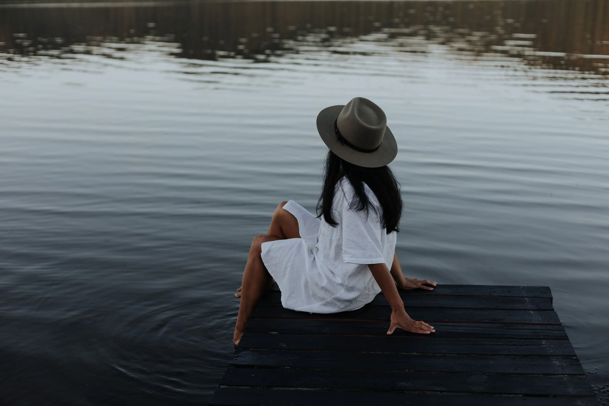 A woman sitting on a dock by the water, wearing a white dress and a wide-brimmed hat, looking out at the lake.