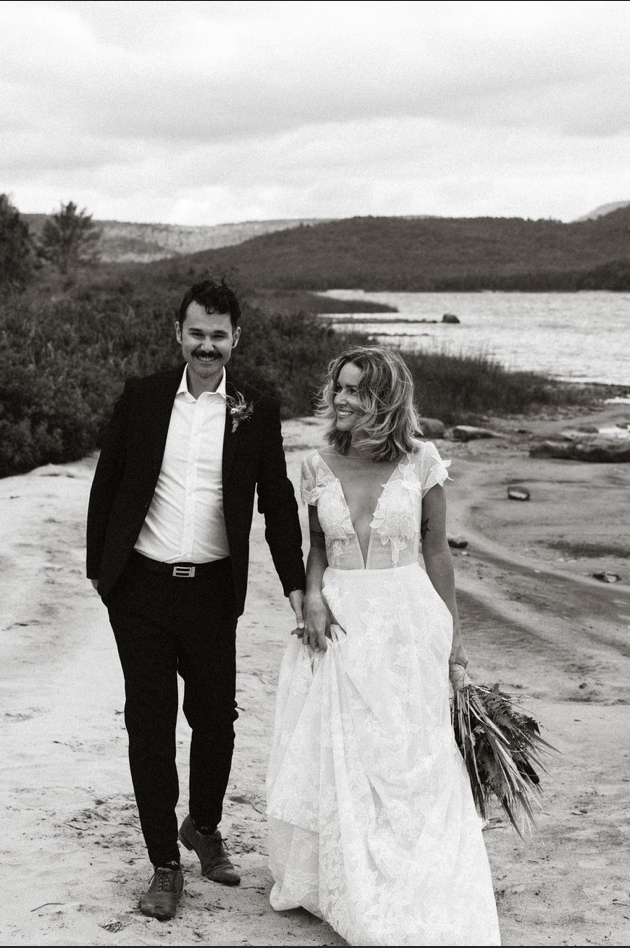 Black and white photo of a smiling couple dressed in wedding attire walking on a beach near a body of water, with mountains in the background.