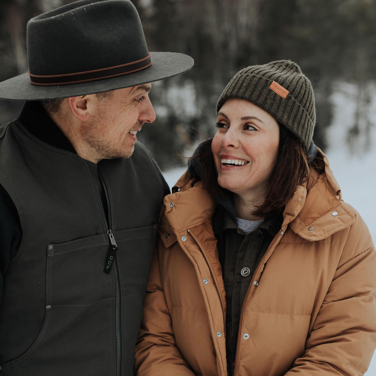 A man and woman smiling and looking at each other outdoors in winter, dressed warmly with jackets and a hat and beanie.